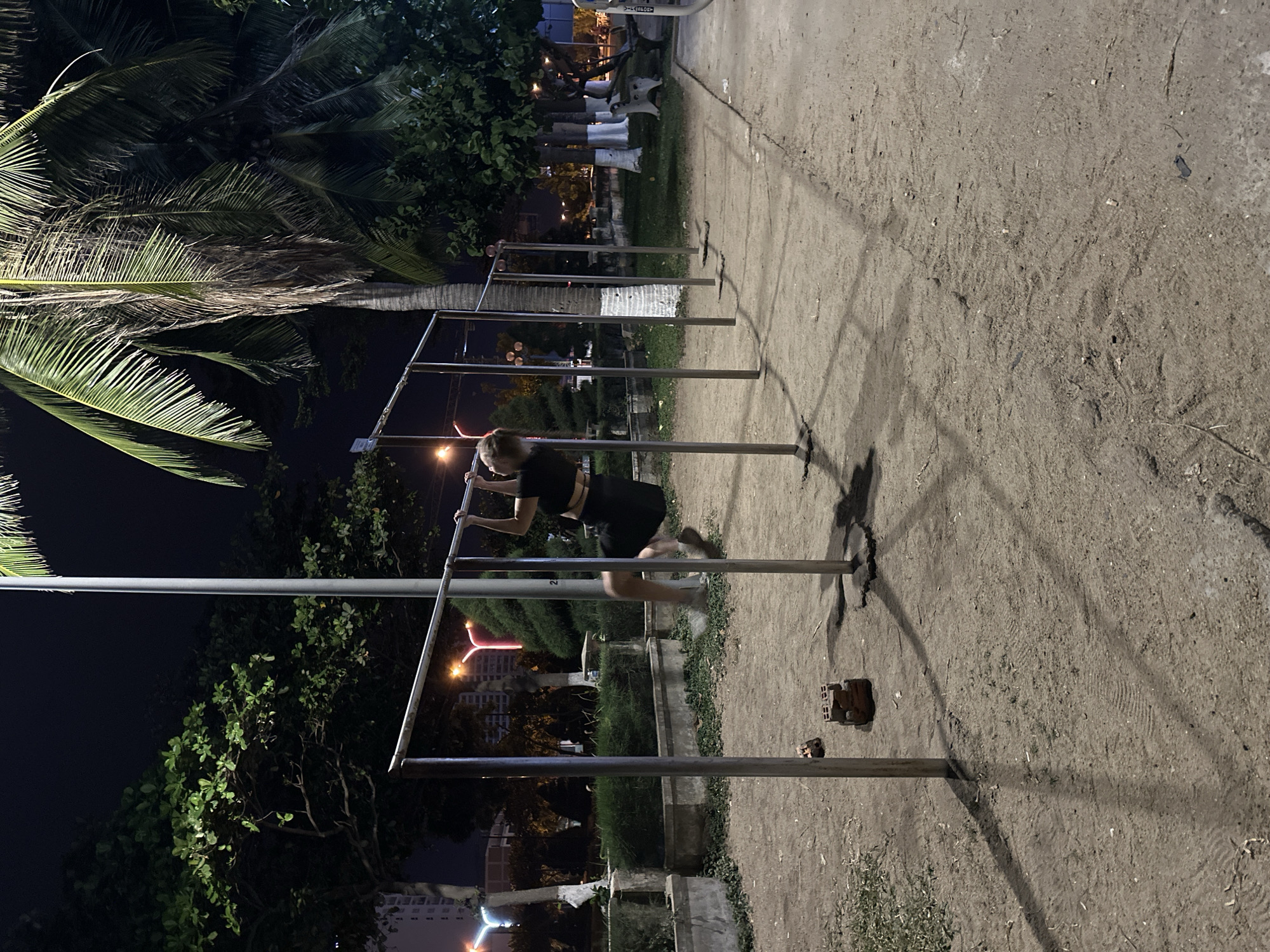 A person is performing pull-ups on a horizontal bar in an outdoor park at night in Nha Trang, Vietnam. The person is wearing dark clothing and is suspended from the bar by their hands. The park features a gravel ground, multiple metal pull-up bars, trees, and palm trees. In the background, buildings and streetlights are visible, indicating an urban environment. There are also two bricks on the ground near one of the pull-up bars. The lighting suggests the scene is illuminated by artificial sources.