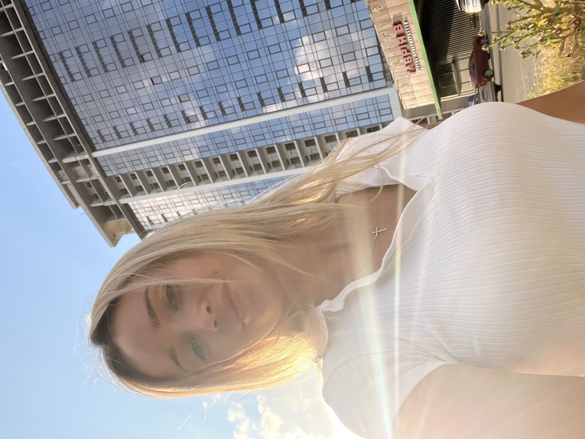 A blonde woman wearing a white short-sleeved ribbed polo shirt and a silver cross necklace is posing for a selfie in front of a modern glass-fronted building. The building is under construction, with concrete beams and unfinished floors visible on the upper levels. The lower facade of the building features a green awning and signage that reads "ГАВРІЯВ" in large red letters, followed by smaller text below. A few cars are parked on the street in front of the building, and some greenery is visible in the foreground. The sky is blue with scattered clouds, and the sun is casting a lens flare across the image, highlighting the woman's hair and shoulder. The location is identified as Fontanka, Ukraine.