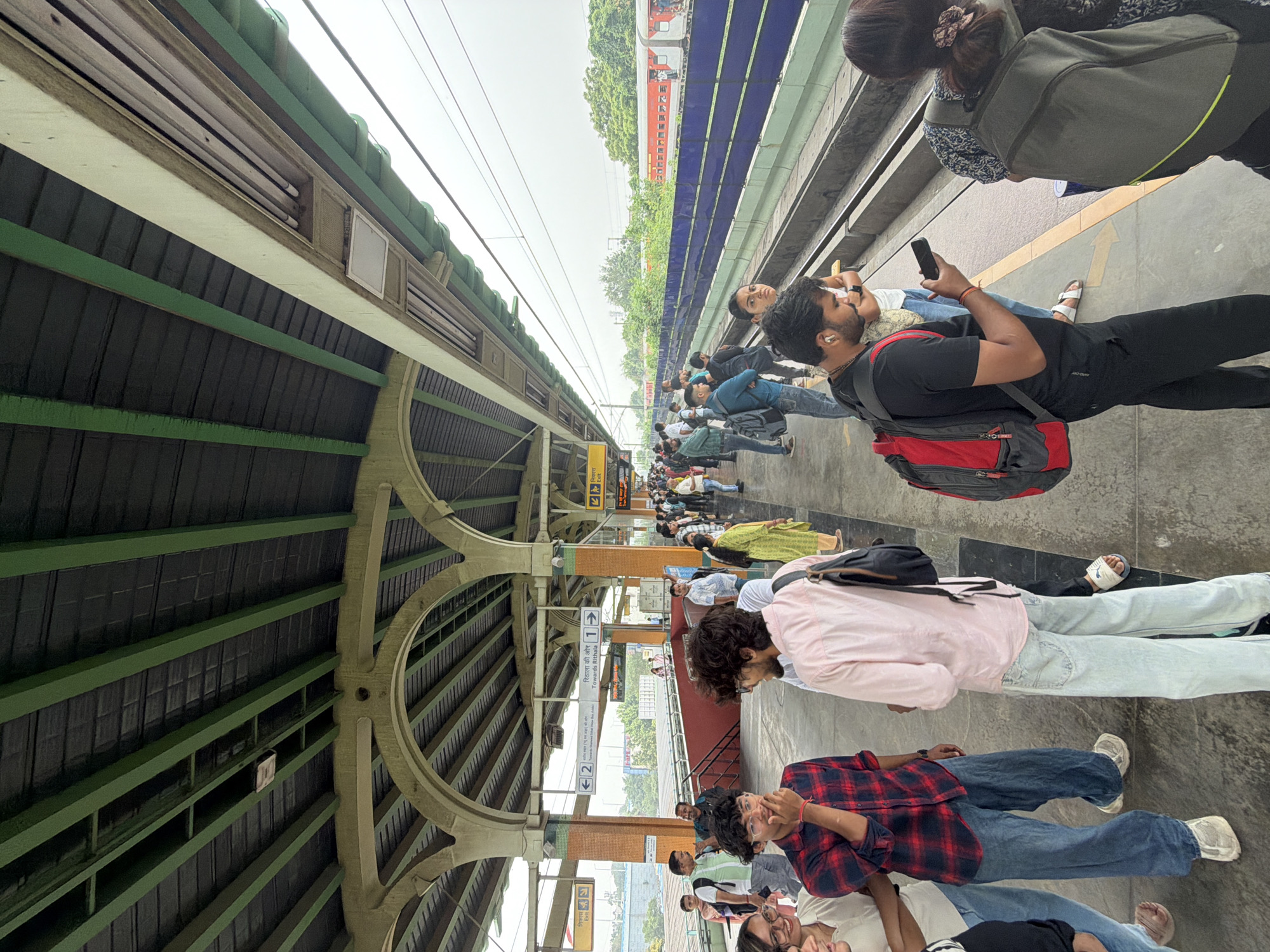 This is a photograph of a metro station platform in Delhi, India. People are waiting on the platform, some are looking at their phones, and others are talking to each other. A train is visible in the background. The platform is covered by a roof supported by pillars. There are signs indicating directions to different platforms. The environment is somewhat overcast.