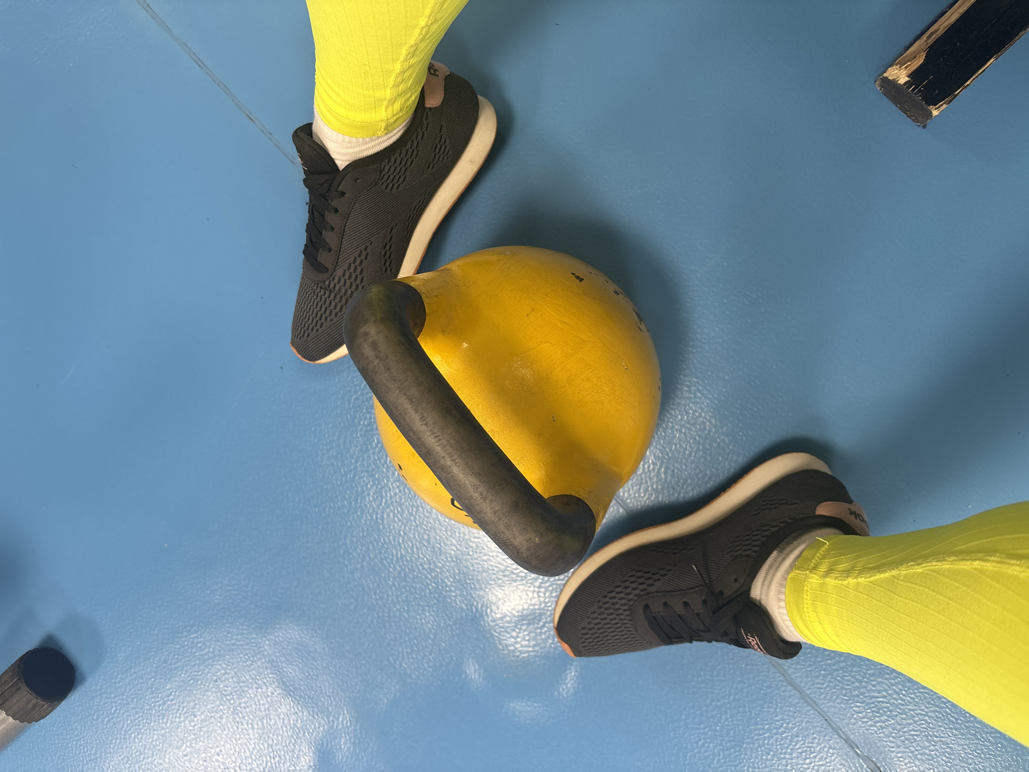 A high-angle, close-up photograph shows a yellow kettlebell on a blue floor, likely in a gym or exercise area. Two pairs of legs and feet are positioned around the kettlebell. Each person is wearing black athletic shoes with white soles, white socks, and bright yellow leggings with a ribbed texture. There's a partially visible black object in the upper right corner and a cylindrical black object in the lower left corner. There is no information available in this image to determine the location in the city of Chaikovsky, Russia.