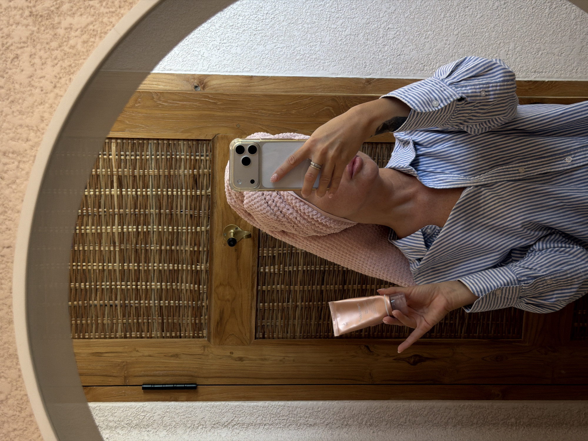 A woman is taking a mirror selfie in what appears to be a bathroom in Mengwi, Indonesia. She is wearing a pink towel wrapped around her head and a blue and white striped button-down shirt. She is holding a tube of cosmetic product in her left hand and her phone in her right hand, with which she is taking the photo. The background shows a wooden door with woven panels and a textured wall.