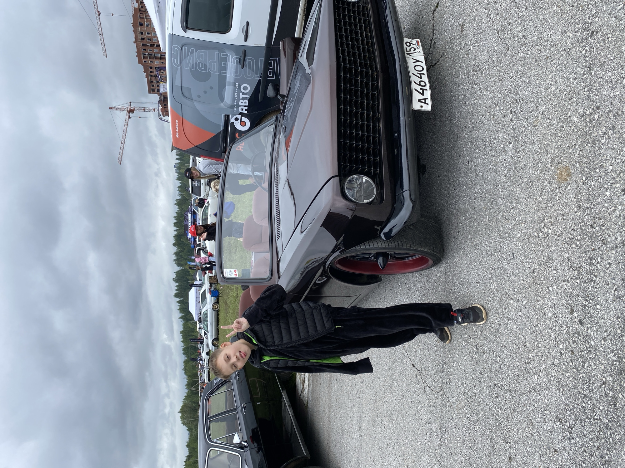 A young Caucasian boy, with light brown hair, wearing a black puffer vest over a black hooded tracksuit with a neon green interior and black sneakers, stands on asphalt in front of a dark brown convertible. He is making a peace sign with his right hand and sticking out his tongue. The convertible has a black grille, a round headlight, and red-rimmed wheels. A license plate on the front reads "A 4640 Y 159". In the background, other cars are visible, along with white tents, trees, and construction cranes against a cloudy sky. The event appears to be an outdoor car show or gathering. The license plate and the inscription on the white van "СЕРВИС АВТО" indicate a likely location in Perm, Russia.