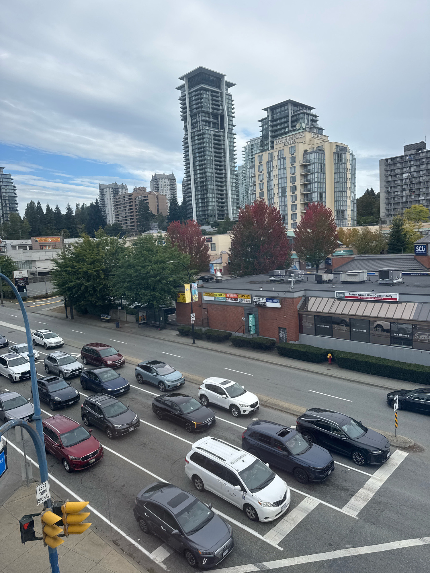 This image captures a traffic scene on a city street in Burnaby, Canada, likely on a cloudy day. The foreground features a traffic light with multiple yellow signals and a "LEFT TURN SIGNAL" sign mounted on a blue pole. Several vehicles are stopped in traffic lanes, including a dark gray SUV, a red SUV, a white minivan, and a dark blue SUV. Beyond the immediate traffic, a multi-lane road continues with more cars.
In the mid-ground, a row of commercial buildings is visible. One building has a sign for "Sutton Group West Coast Realty" and "SCU." Another building appears to be a hotel with a sign that reads "EXECUTIVE PLAZA HOTEL." Trees with red and green foliage are interspersed among the buildings.
The background is dominated by several tall, modern high-rise residential buildings. The sky is overcast with a mix of gray and lighter clouds. The overall impression is of an urban environment with significant vehicle and building density.
