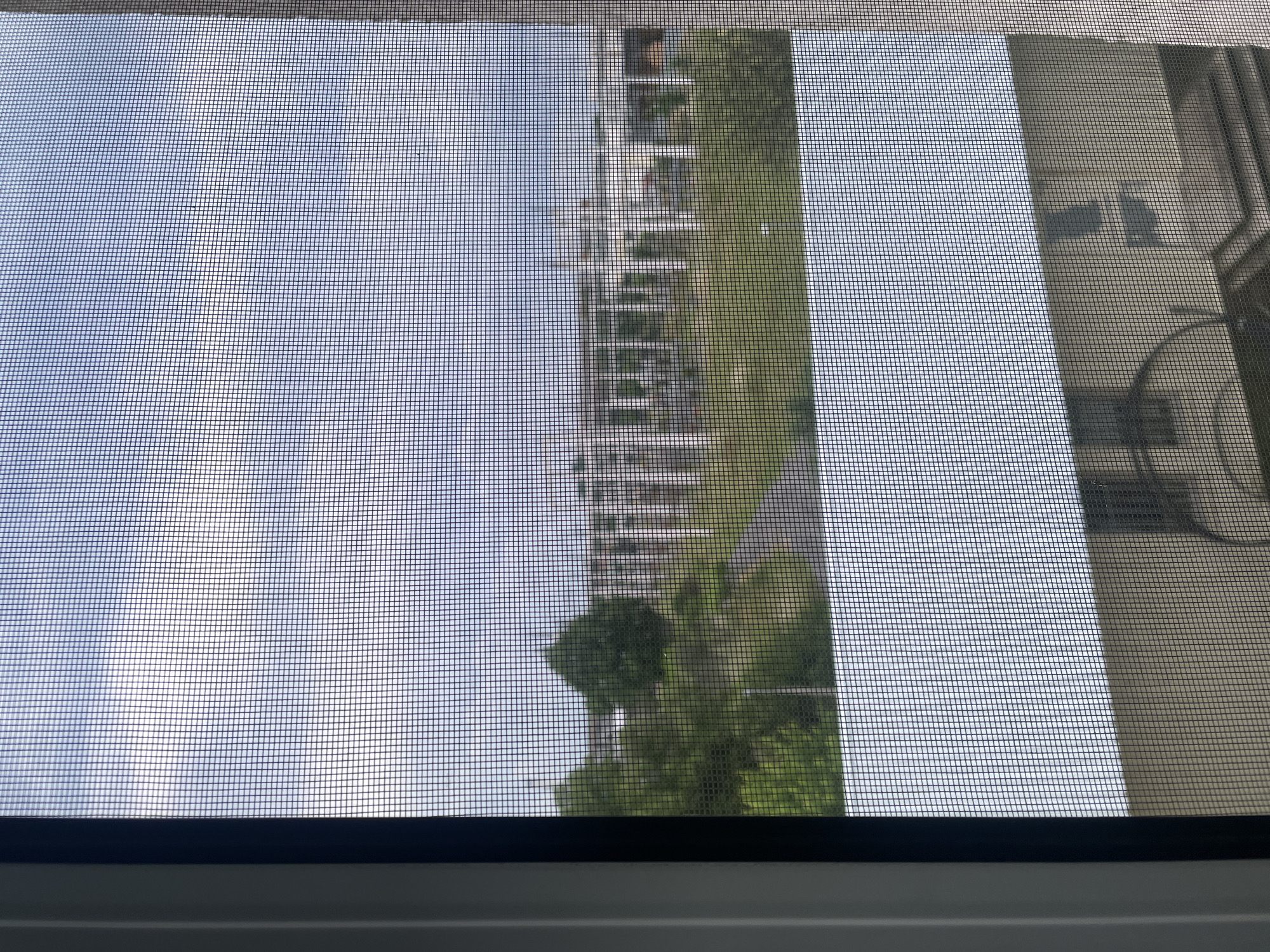 The image displays an outdoor scene viewed through a fine-mesh window screen, which creates a prominent grid pattern across the entire frame. The image appears to be rotated approximately 90 degrees clockwise from its natural orientation.
On the left side of the frame, a bright, light blue sky with wispy white clouds is visible through the screen. Moving towards the center, a modern, multi-story building with numerous balconies or ledges appears. This building is light in color, possibly white or light grey, and stretches horizontally across the middle of the frame due to the image's rotation. To the right of this building, a dense area of vibrant green foliage, likely trees, fills a significant portion of the view. Further to the right, another building, darker in tone and partially obscured by the screen, can be seen, exhibiting architectural details like windows and potentially a curved structure.
The bottom edge of the image is defined by a dark, horizontal window frame, below which a wider, lighter-colored section of the window sill or frame is visible.
The scene depicts a daytime view under clear or partly cloudy weather conditions, suggesting an urban or suburban environment with a mix of buildings and greenery. There are no people or discernible activities taking place. No visible text or strong emotions are conveyed by the image.