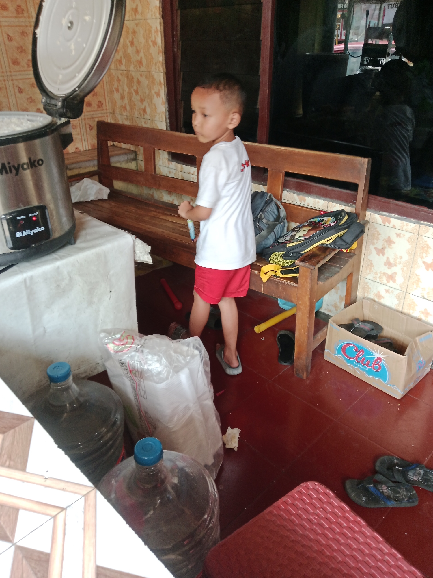 The image depicts a young boy wearing a white t-shirt and red shorts, standing in an indoor area with tiled floors and patterned walls. He is looking towards the right side of the frame. In the background, there is a wooden bench with a backpack placed on it. To the left, a rice cooker with an open lid is visible on a white-covered surface, next to large plastic water bottles. A cardboard box filled with flip-flops is located to the right of the bench. The scene appears to be in a residential setting, possibly a home or a small shop, in Kediri City, Indonesia. The lighting suggests it is daytime.