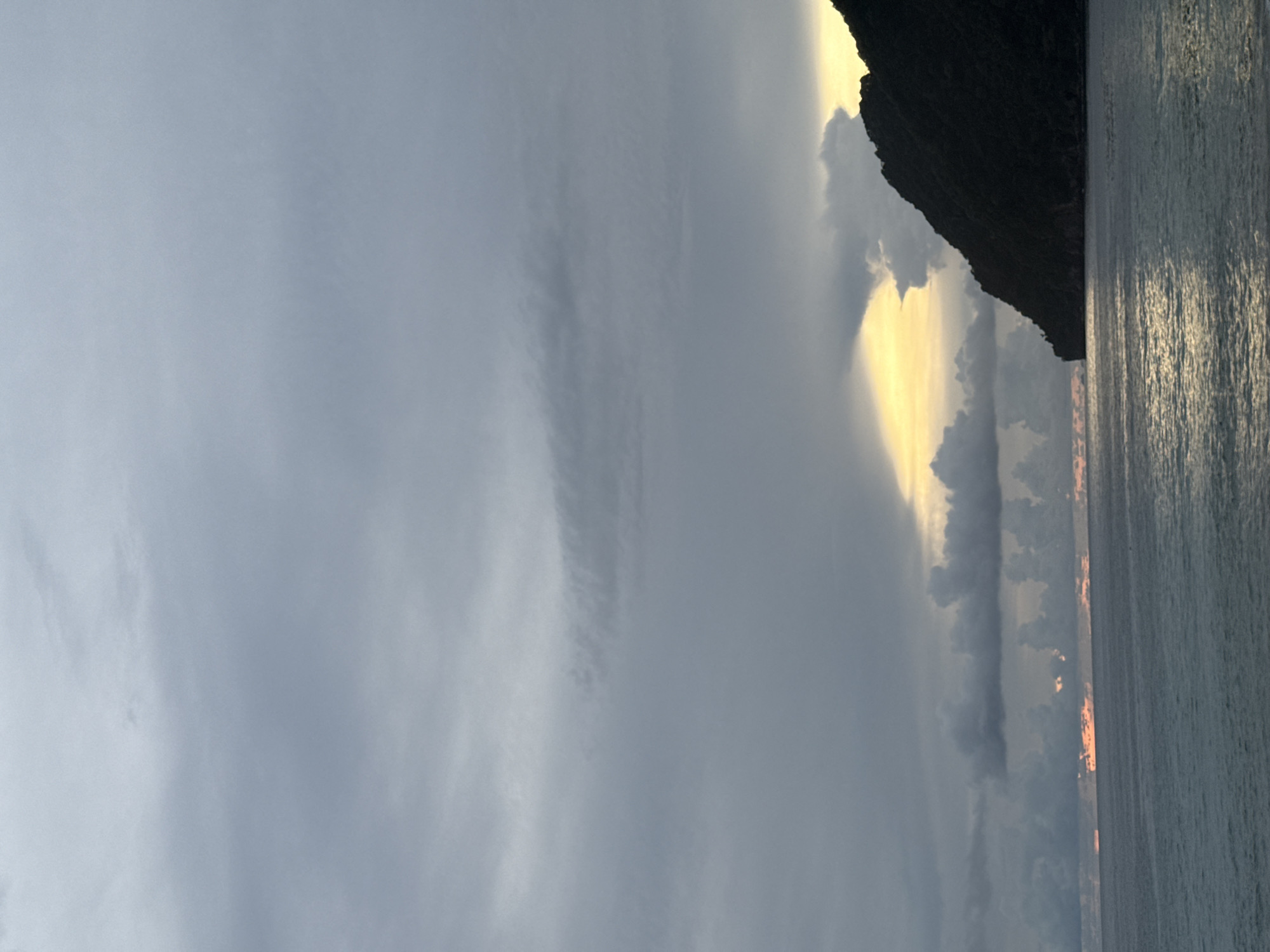 This is a photograph of the sea and a mountainous coastline at dusk. The sky is filled with dark, heavy clouds, with a bright yellow glow near the horizon where the sun is setting behind the mountain. The sea is a deep gray-blue, with gentle waves reflecting the light from the sky. A dark, rugged mountain juts out from the right side of the frame, its silhouette stark against the colorful sky. The location is Meunasahbale, Indonesia.