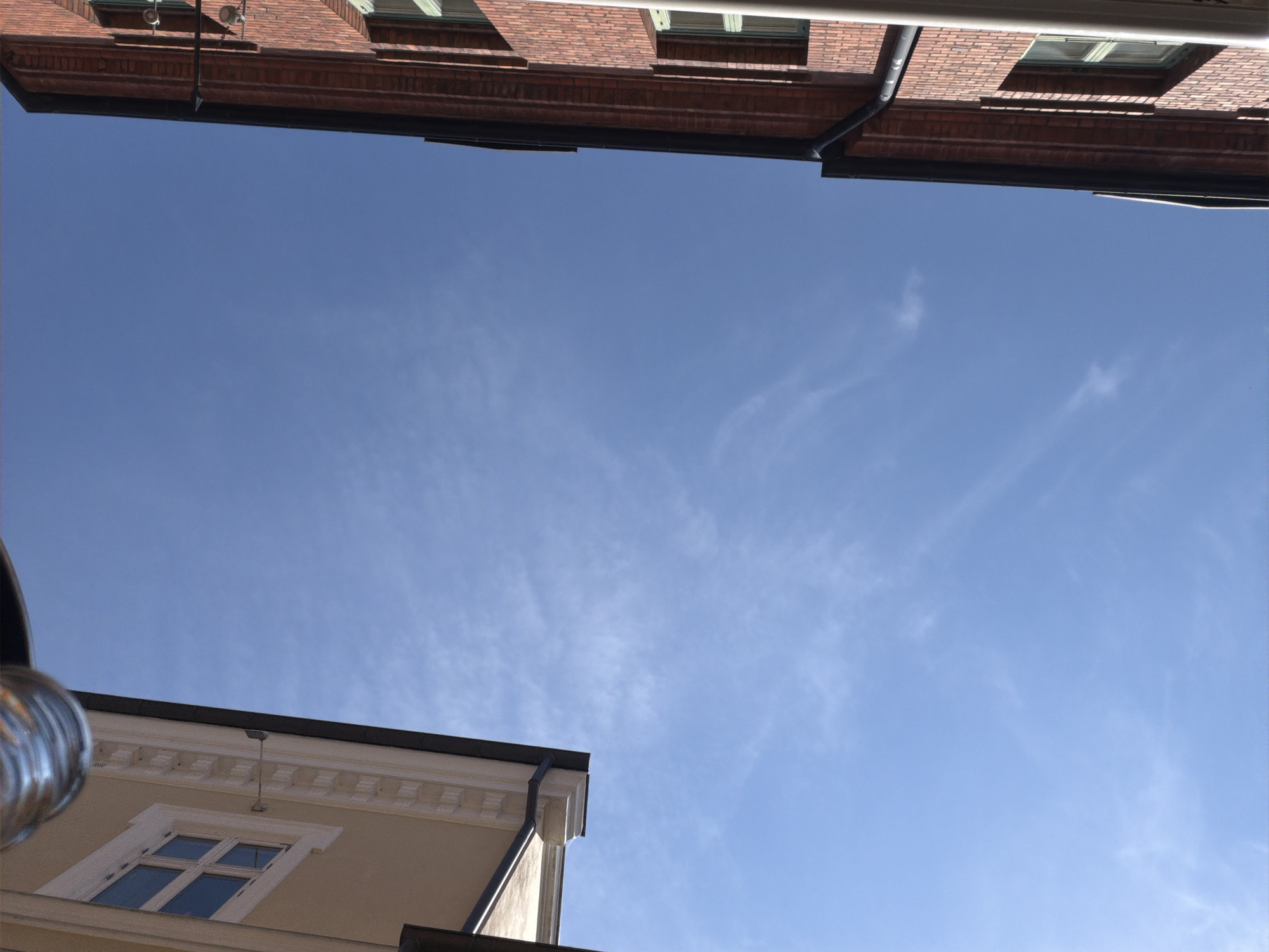 The image is an upward-facing shot taken between two buildings, one on the left with a light-colored facade and ornate trim, and another on the right constructed of red brick. The sky is a clear blue with faint, wispy clouds. A metal pipe or gutter is visible at the very top of the frame, to the left. The buildings appear to be of a traditional architectural style, possibly in an urban setting. The perspective suggests the photographer is standing in a narrow alley or street. Without further context or identifying landmarks, it is impossible to definitively place this image within Malmö, Sweden.
