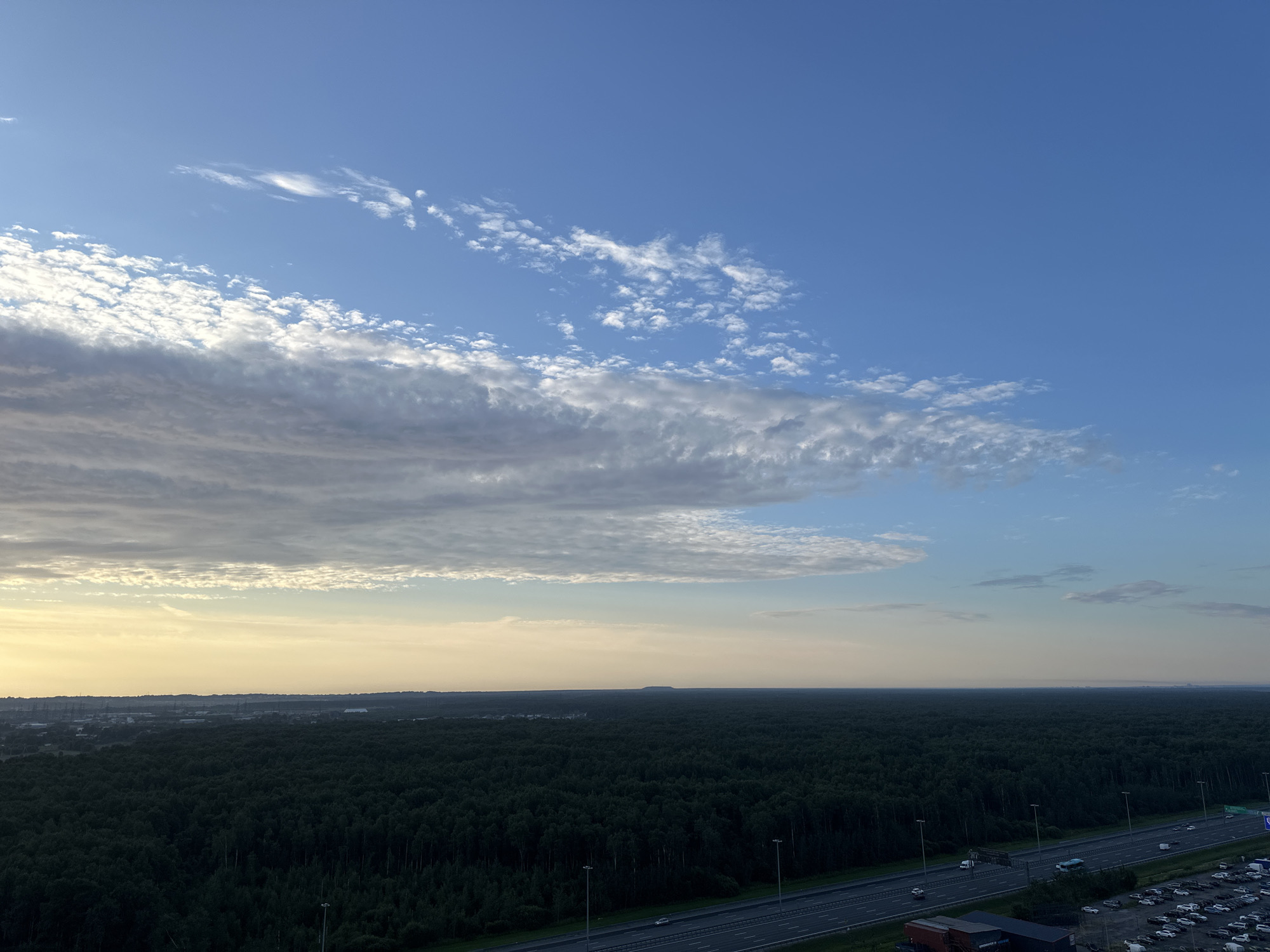 An aerial, wide-angle photograph captures a vast expanse of dark green forest beneath a bright blue sky with scattered, wispy white clouds. A multi-lane highway runs diagonally across the lower right portion of the frame, with vehicles visible on its surface and a parking lot full of cars to its right. A faint, hazy horizon stretches across the scene, with a very subtle rise in elevation visible in the distance. The light suggests it is either early morning or late evening, casting a soft glow on the sky.