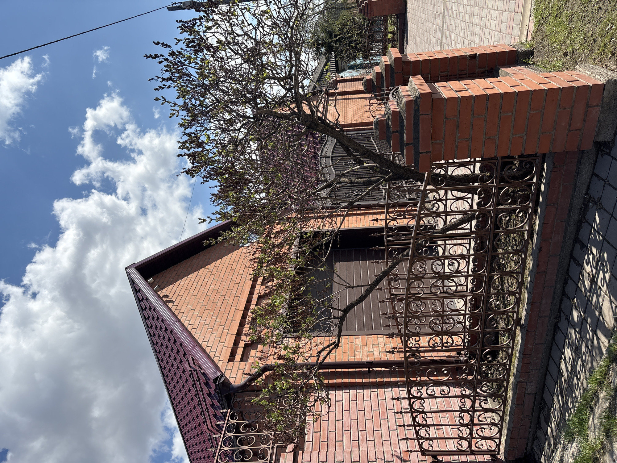 The image shows a red brick building with a dark-colored roof under a partly cloudy blue sky. A wrought iron fence runs along the side of the building, with a tree in front of it. A brick pathway is visible near the fence, with grass growing nearby. The scene appears to be an exterior shot of a residential or commercial property.