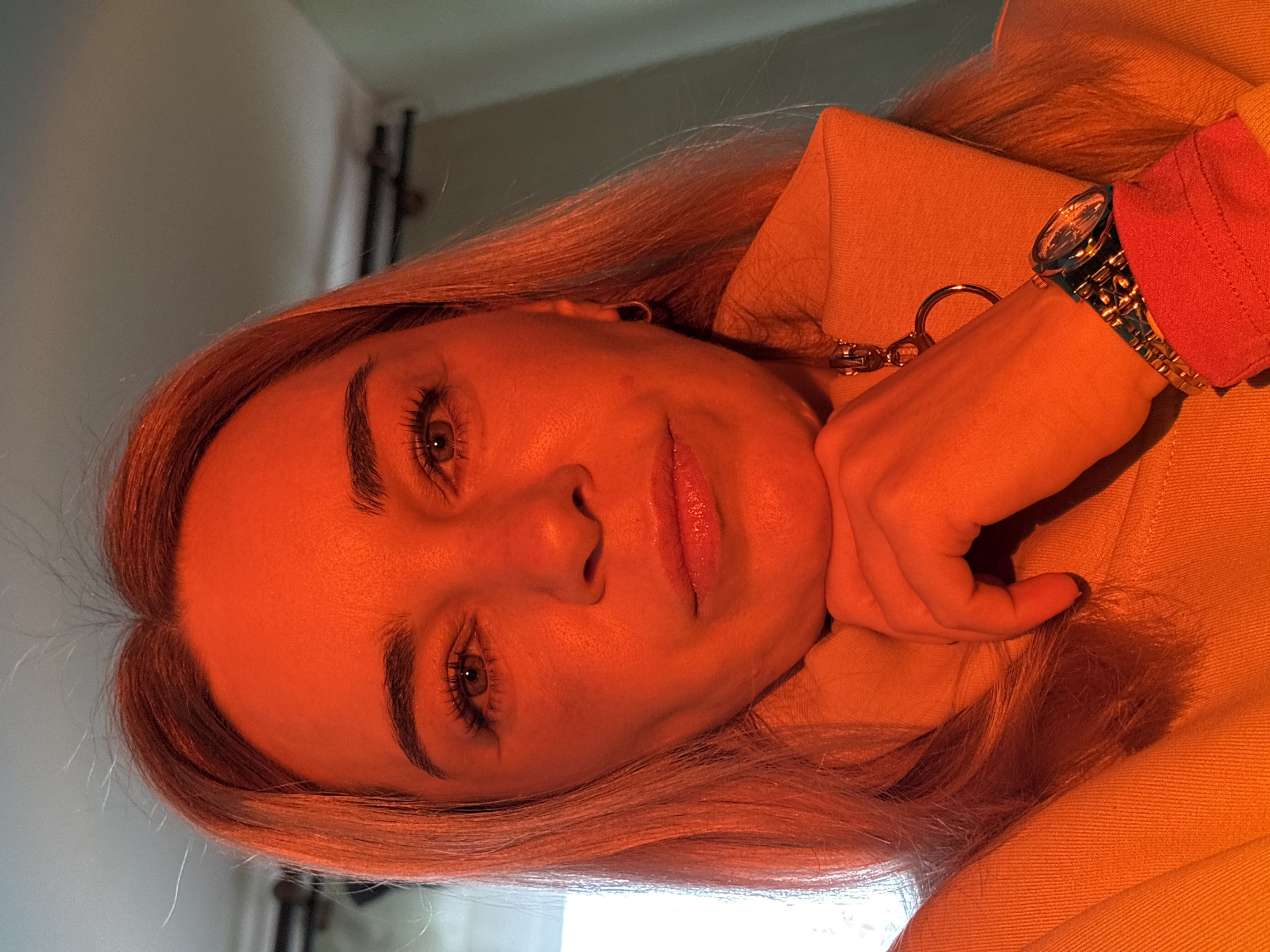 A woman is indoors. She has fair skin, light-colored hair, and is wearing a watch on her left wrist. Her hand is resting on a tan jacket. There is a blurred background with a window and what appears to be a curtain rod. The location is in the city of Unknown, Unknown.