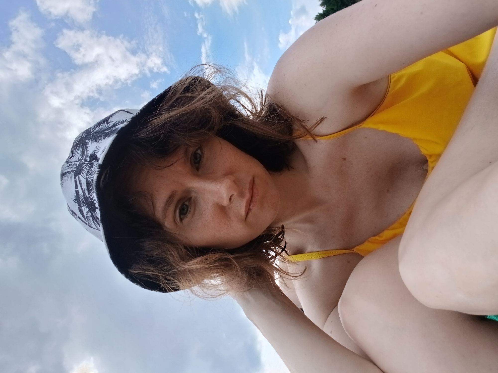 A woman with shoulder-length brown hair and freckles is wearing a bucket hat with a palm tree pattern and a yellow swimsuit. She is looking at the camera with a neutral expression, her left arm bent and her hand resting on her head. The background is a cloudy blue sky, with trees visible on the right side. The photograph was taken outdoors, possibly near a body of water, given the relaxed attire and the natural setting. It is not possible to determine the specific location in Revda, Russia, from the image alone.