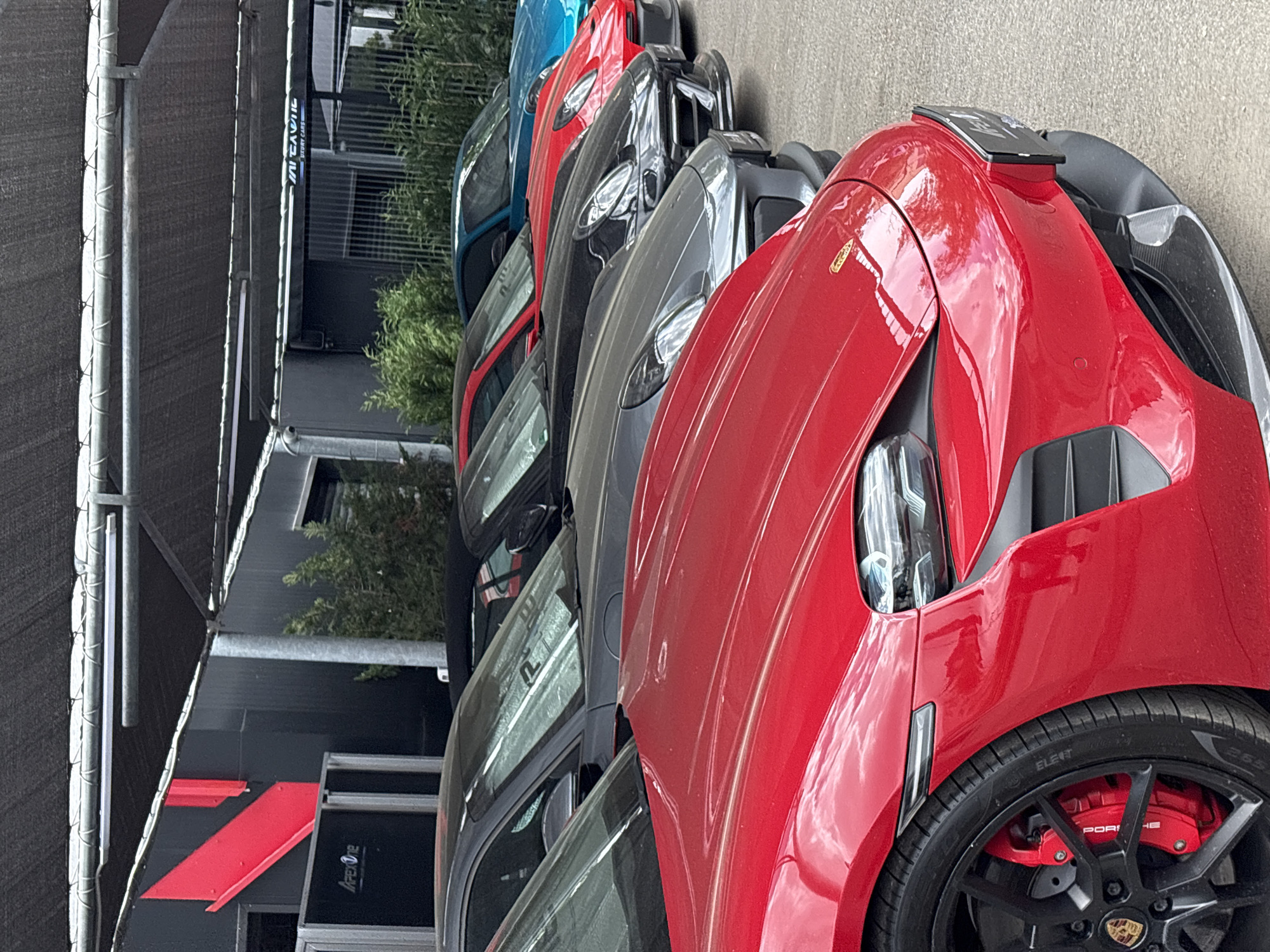 The image captures a lineup of luxury vehicles in front of a building with the "IMPERIO" logo, located in Sofia, Bulgaria. The lineup includes several high-end cars in shades of red, gray, black, and blue. The vehicles are tightly packed, likely in a display setting at a dealership. The closest car, predominantly red, features black wheels with red brake calipers and a Porsche logo. The building behind the cars has a modern design, with a black exterior accented by a red stripe. There is greenery partially visible between the building and the cars. The ground appears to be paved.