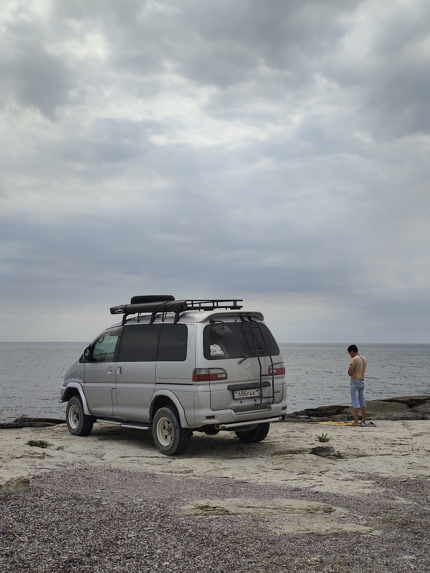 A silver off-road van is parked on a rocky shore near the Caspian Sea in Aqshuqyr, Kazakhstan. A young man, wearing a light brown t-shirt and blue shorts, stands facing away from the van, looking down at a yellow object on the ground. The van is equipped with a roof rack and a spare tire mounted on the back. The sky is overcast with gray clouds. The ground is covered in small pebbles and rocks.