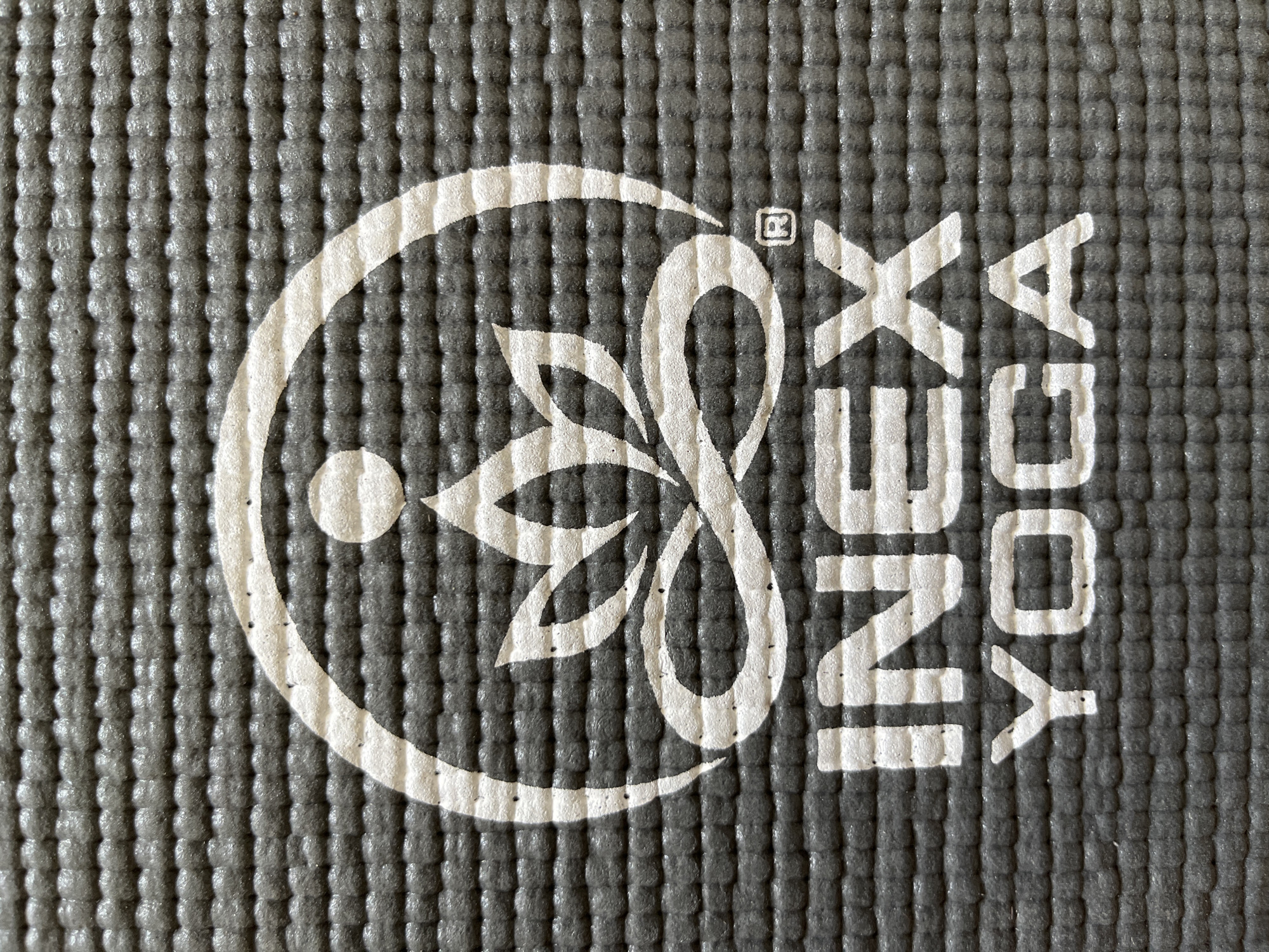 A close-up, overhead shot shows a gray yoga mat with a white logo. The logo is a stylized lotus flower within a circle, with the letters "INEX YOGA" printed below it, as well as a registered trademark symbol to the right of the lotus. The mat's surface has a textured, pebbled pattern.