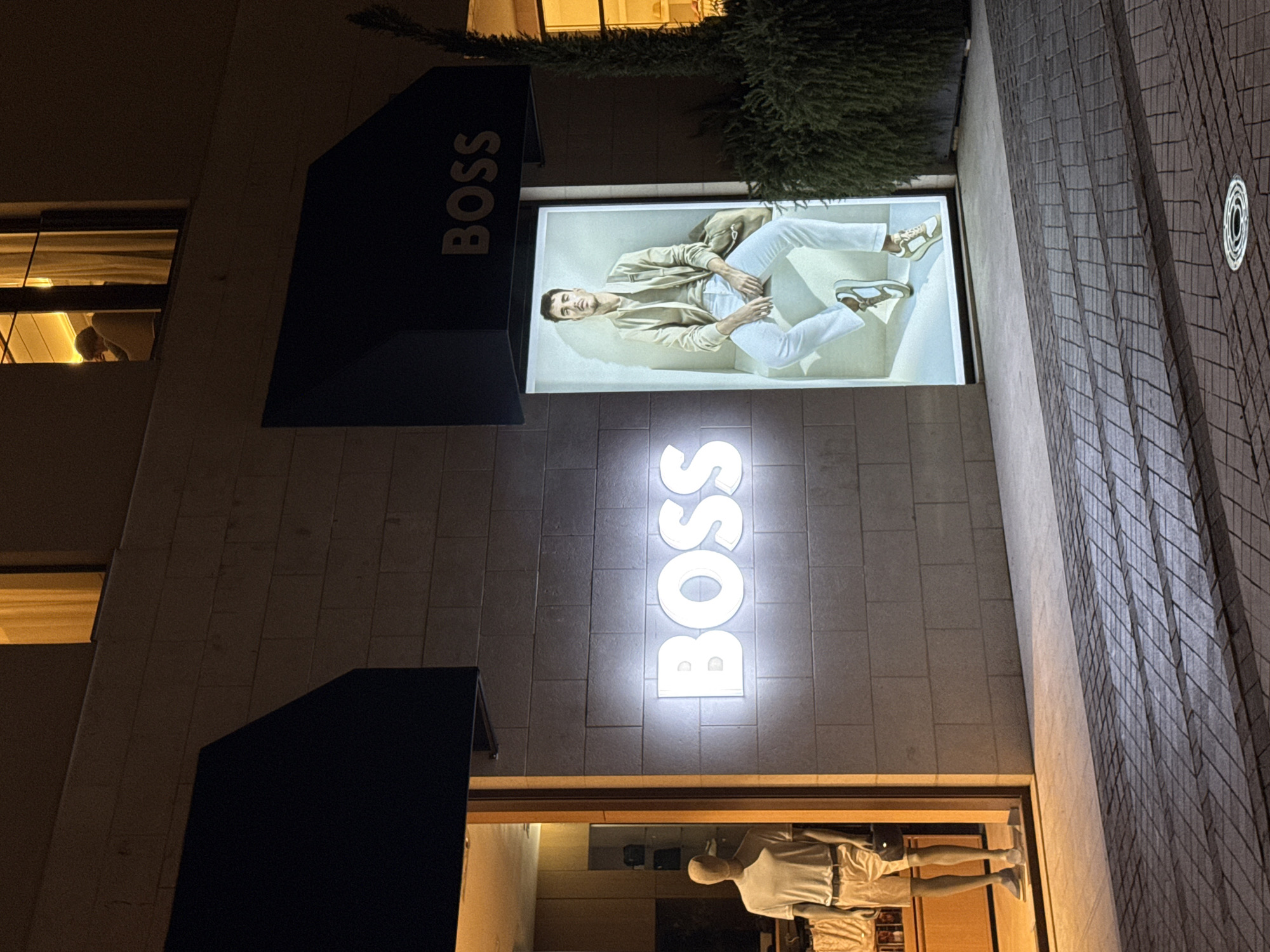 The image shows the exterior of a BOSS store at night in Tivat, Montenegro. A mannequin wearing a white t-shirt and shorts stands in a lit storefront on the left. To the right of the storefront, a large, illuminated sign with the word "BOSS" in white neon letters is mounted on a brick wall. Further to the right, a large, illuminated advertisement displays a man in casual clothing sitting down. A dark green, dense bush is positioned to the right of the advertisement. Above the storefront and advertisement, dark awnings are visible. The ground in front of the store is paved with interlocking bricks, and the lighting casts distinct shadows. In the upper right portion of the image, a window with warm yellow light is visible, with a person faintly silhouetted inside.