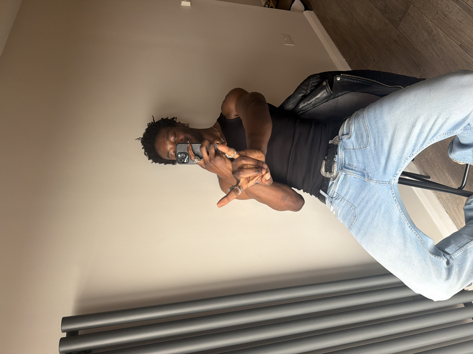 A medium indoor shot shows a Black person using a mobile phone to take a photo in a room with beige-colored walls. The person is wearing a black tank top, light blue denim jeans with a decorative belt buckle, and a black leather jacket is draped over the back of the chair on which the individual is sitting. The flooring is a wood grain pattern. A gray radiator is visible at the bottom of the frame. The location of the scene cannot be determined from the image.