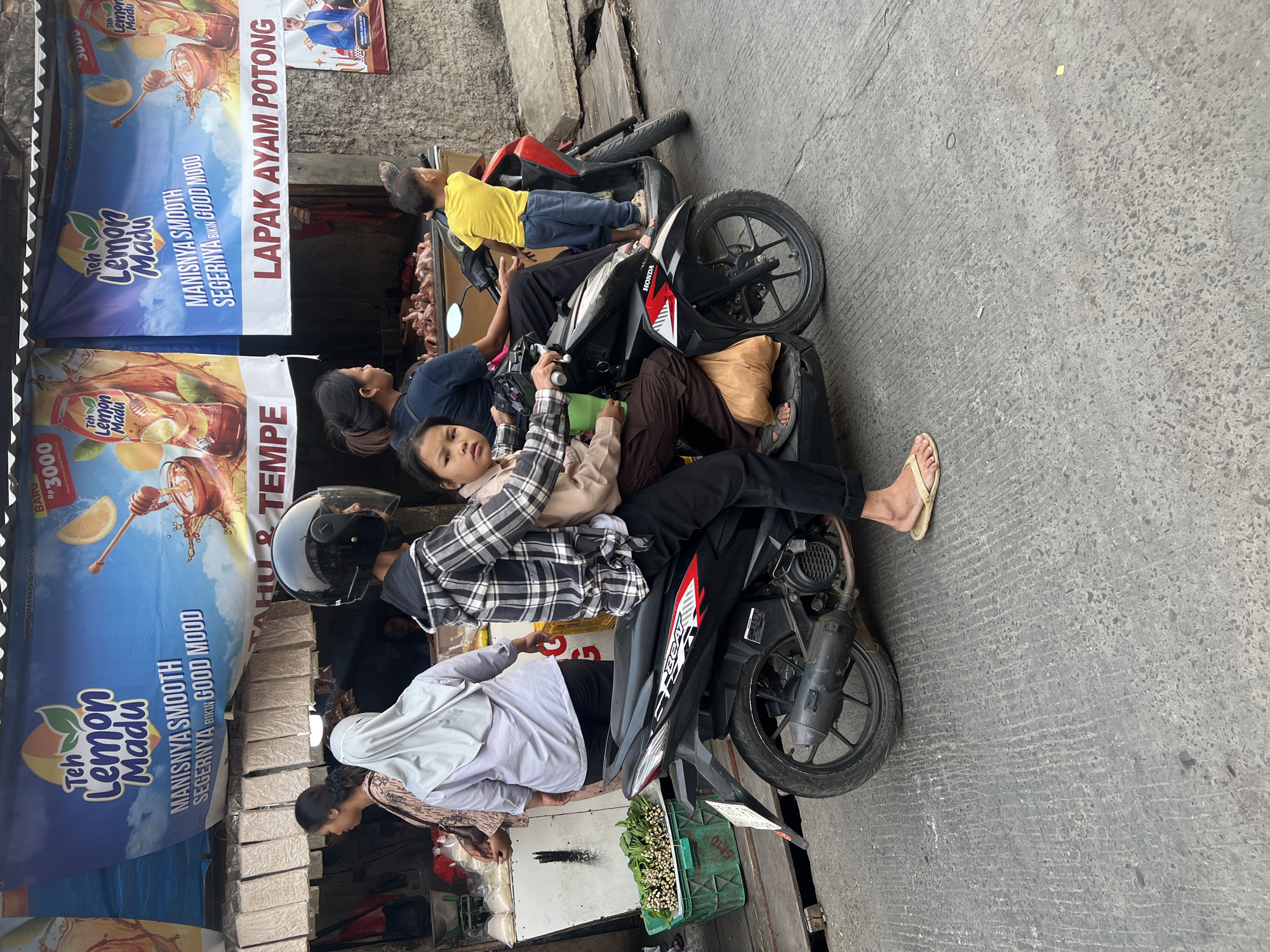 The image captures a street scene in Citeureup, Indonesia, featuring a busy market environment. In the foreground, a family of four is on a black Honda Beat motorcycle. The driver, wearing a black helmet and a plaid jacket, is in the front. Behind them, a young girl sits between the driver and a woman. Another young boy, in a yellow shirt and blue pants, stands on the back of a red scooter parked next to them.
The background reveals market stalls with various goods. Banners advertising "Teh Lemon Madu" (Lemon Honey Tea) hang above the stalls. One banner prominently displays "MANISNYA SMOOTH SEGERNYA BIKIN GOOD MOOD" (Sweetly Smooth, Refreshingly Makes a Good Mood). Another banner reads "LAPAK AYAM POTONG" (Cut Chicken Stall). Visible goods include bundles of green vegetables in a green crate, and what appear to be packaged food items like tempeh hanging from the stalls. The overall impression is one of a vibrant, everyday marketplace.