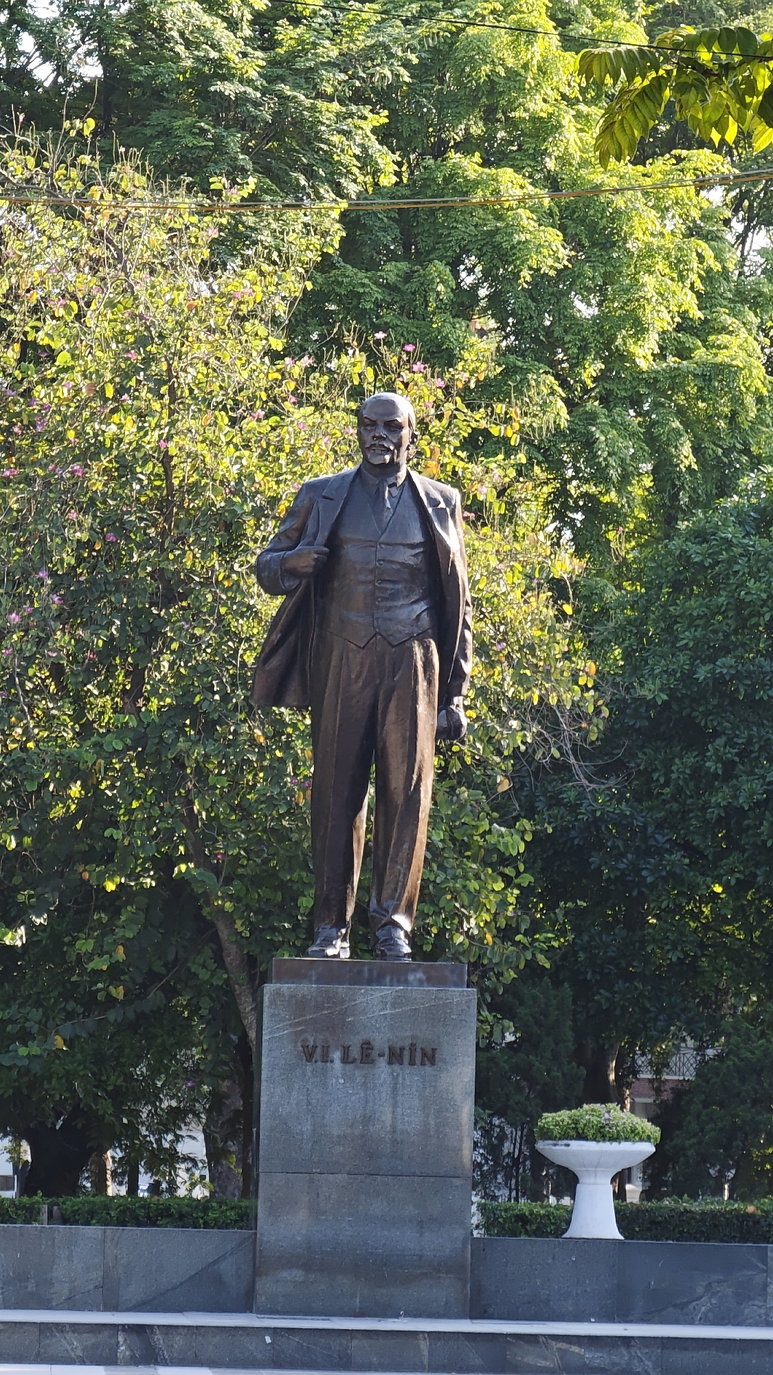 The image depicts a bronze statue of Vladimir Lenin standing on a gray stone pedestal in a park-like setting. The statue is of a man in a suit with an open jacket, one hand inside the jacket, and the other hand at his side. The pedestal bears the inscription "VL LE-NIN." Behind the statue are various trees and bushes, some with flowers. To the right of the statue, on a lower platform, is a white planter containing green plants. The location is reportedly in Hà Nội, Vietnam.