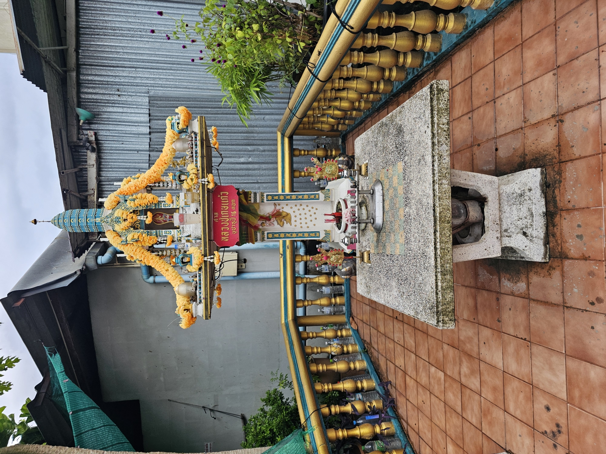 An outdoor spirit house, or San Phra Phum, is depicted in this medium shot. The spirit house is ornately decorated with yellow and orange garlands. It is situated on a stone table with a patterned surface, and the table stands on a concrete base. In front of the table, there is a metal stand with red incense sticks burning in a white pot. To the right of the table, there are decorative figurines, and further right, a lush green plant with small purple flowers. The spirit house and table are surrounded by a golden balustrade with blue accents. The ground is tiled with reddish-brown square tiles, showing some wear and tear. In the background, there is a corrugated metal wall and a gray concrete wall, with a green tarp stretched over a sloped roof. The sky is overcast. The scene appears to be in a somewhat aged outdoor area, possibly within a residential or commercial property in Bangkok, Thailand. There are no people visible in the image.