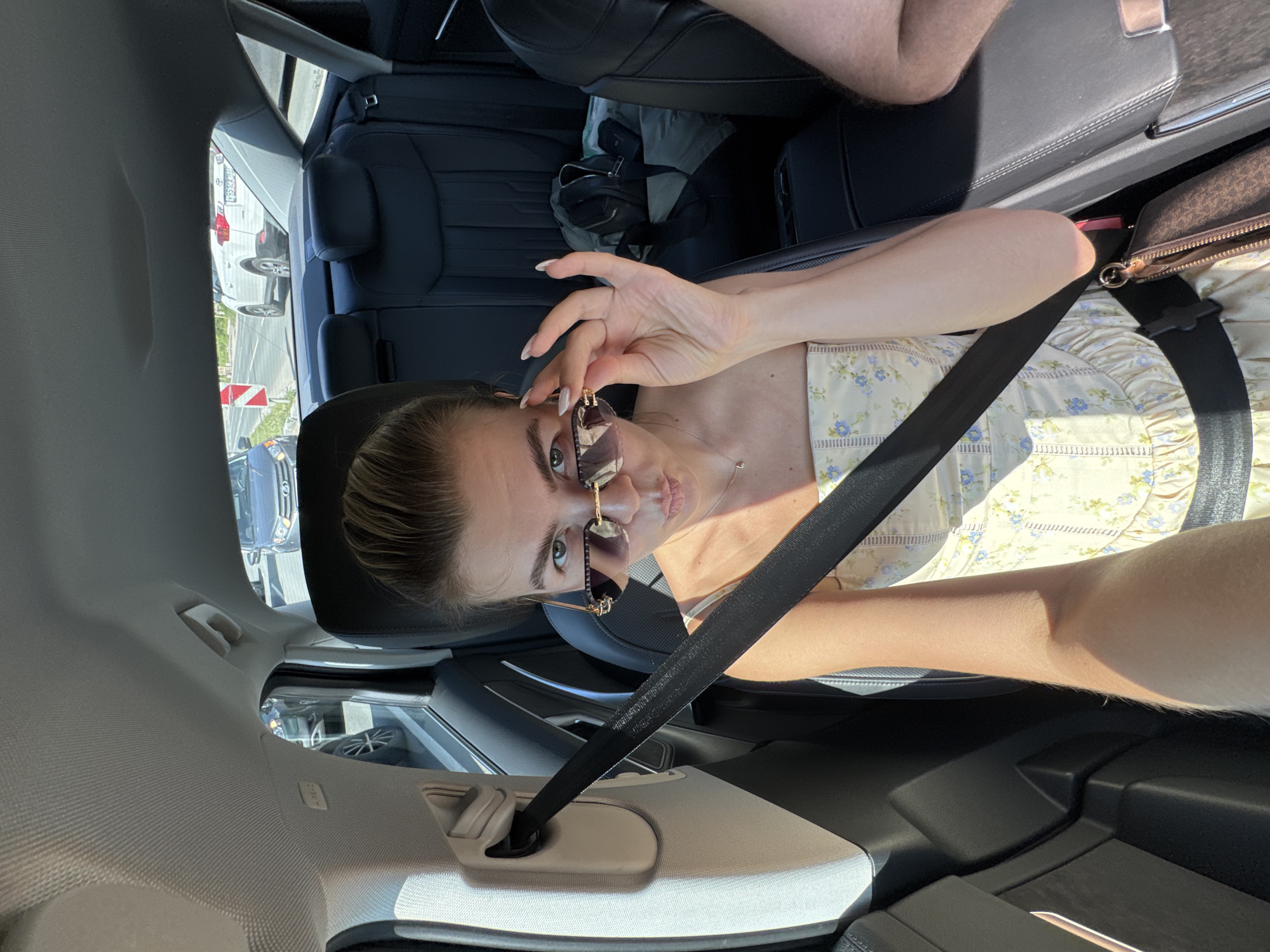 A young woman with blonde hair pulled back is seated in the back seat of a car, wearing a light-colored floral dress and sunglasses. She is holding her sunglasses with her right hand, looking at the camera with her lips pursed. A seatbelt is visible across her chest. The car interior appears to be black leather. Outside the car windows, other vehicles and a roadside scene are visible. The lighting suggests it is daytime. The location is unknown.