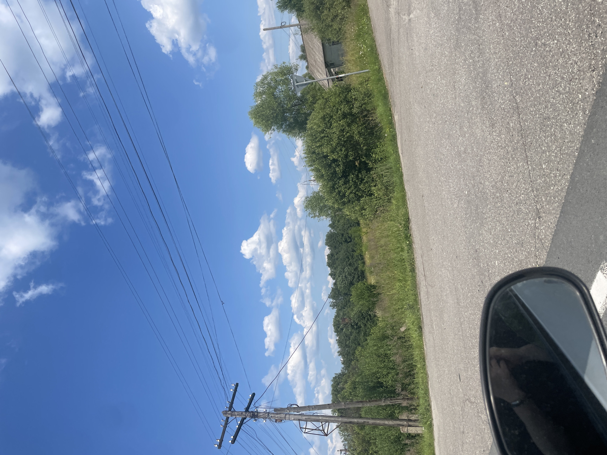 The image depicts a rural roadside scene in the vicinity of Pomogalovo, Russia, under a clear blue sky with scattered white clouds. A gray asphalt road curves slightly through the frame. On the left side of the road, a wooden utility pole with multiple wires is visible, alongside overgrown green vegetation and trees. In the background, a dense line of trees forms a natural boundary, and further to the right, an aged, weathered wooden house with a gray roof stands among the foliage. A side-view mirror of a vehicle is partially visible in the lower left corner, reflecting a person's arm and hand. The overall impression is of a quiet, possibly abandoned, rural setting on a bright, sunny day.
