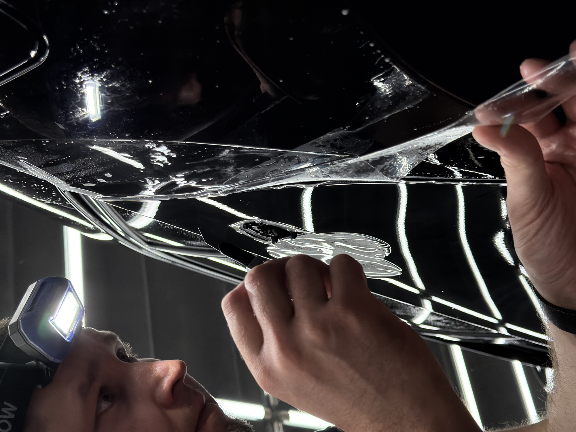 A person with a headlamp is applying a clear protective film to a black surface, likely a vehicle. The film is partially adhered, with bubbles and moisture visible underneath. The person is using a black squeegee-like tool to smooth the film. The location is unknown.