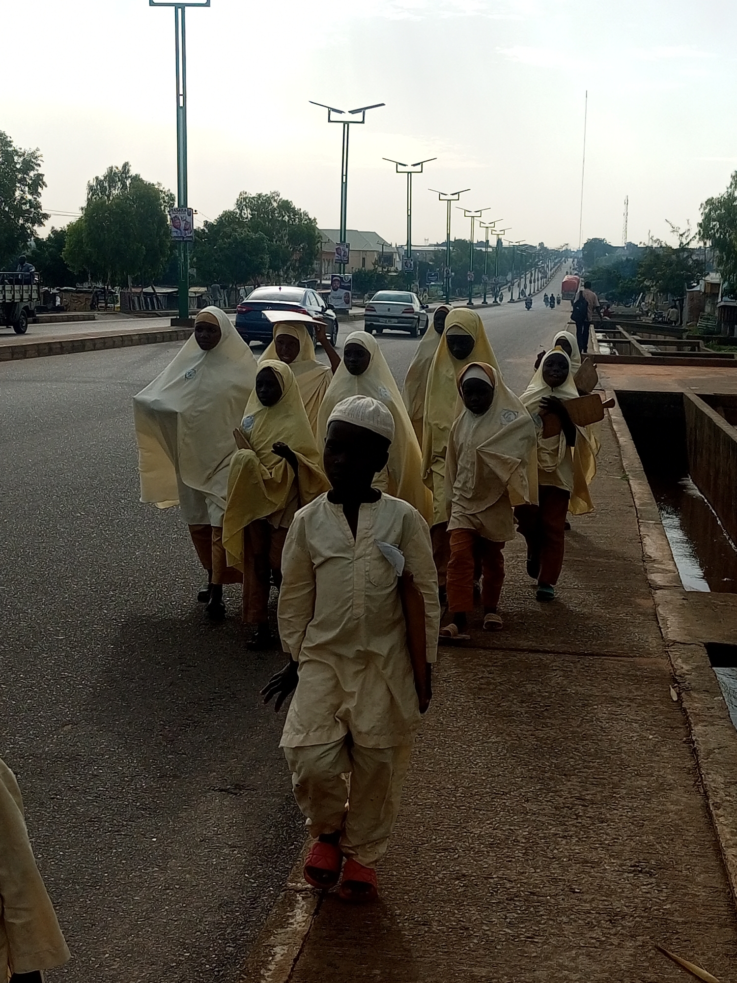 A group of eight children, primarily girls wearing light-colored hijabs and loose-fitting garments, are walking in single file on a paved sidewalk in Wuro Dole, Nigeria. The child in the foreground, a boy wearing a white cap and matching outfit, walks slightly ahead of the group, facing forward. The other children follow behind him, some looking to the side, and one girl carrying a wooden object that resembles a rolling pin. The sidewalk is adjacent to a two-lane road where cars, including a white sedan and a dark SUV, are visible in motion. Beyond the road, trees and buildings line the street. Streetlights with solar panels are positioned along the road. The sky is overcast and appears to be late afternoon or early morning. The environment suggests an urban setting with infrastructure and typical street life.
