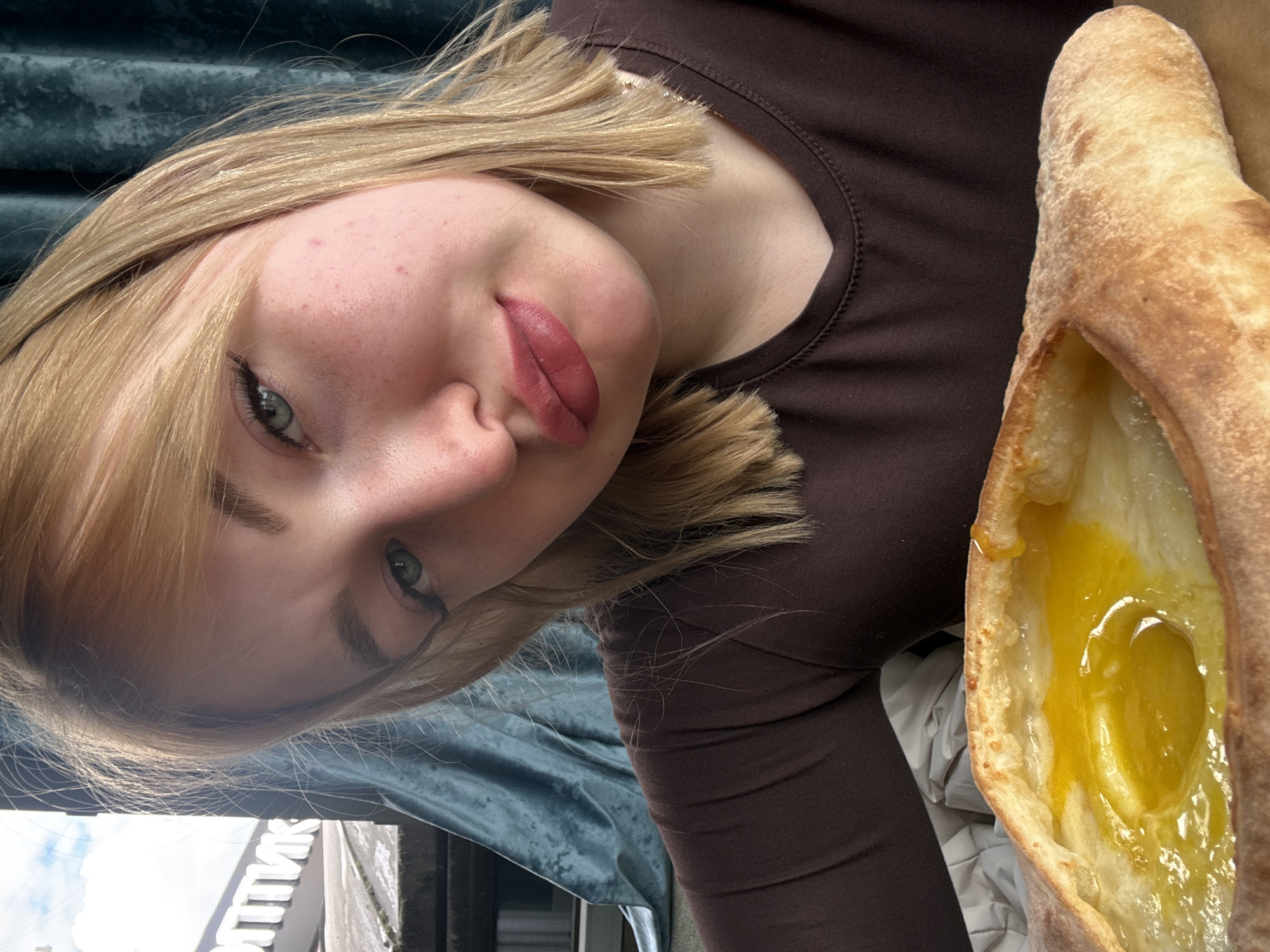 A young woman with blonde hair and makeup is taking a selfie in what appears to be a cafe or restaurant. She is wearing a dark brown long-sleeved shirt and is holding a pastry dish filled with cheese and a bright yellow egg yolk. The background shows a window with a view of a city street and blue curtains. Text visible on a sign outside the window suggests the location is Krasnogorsk, Russia.