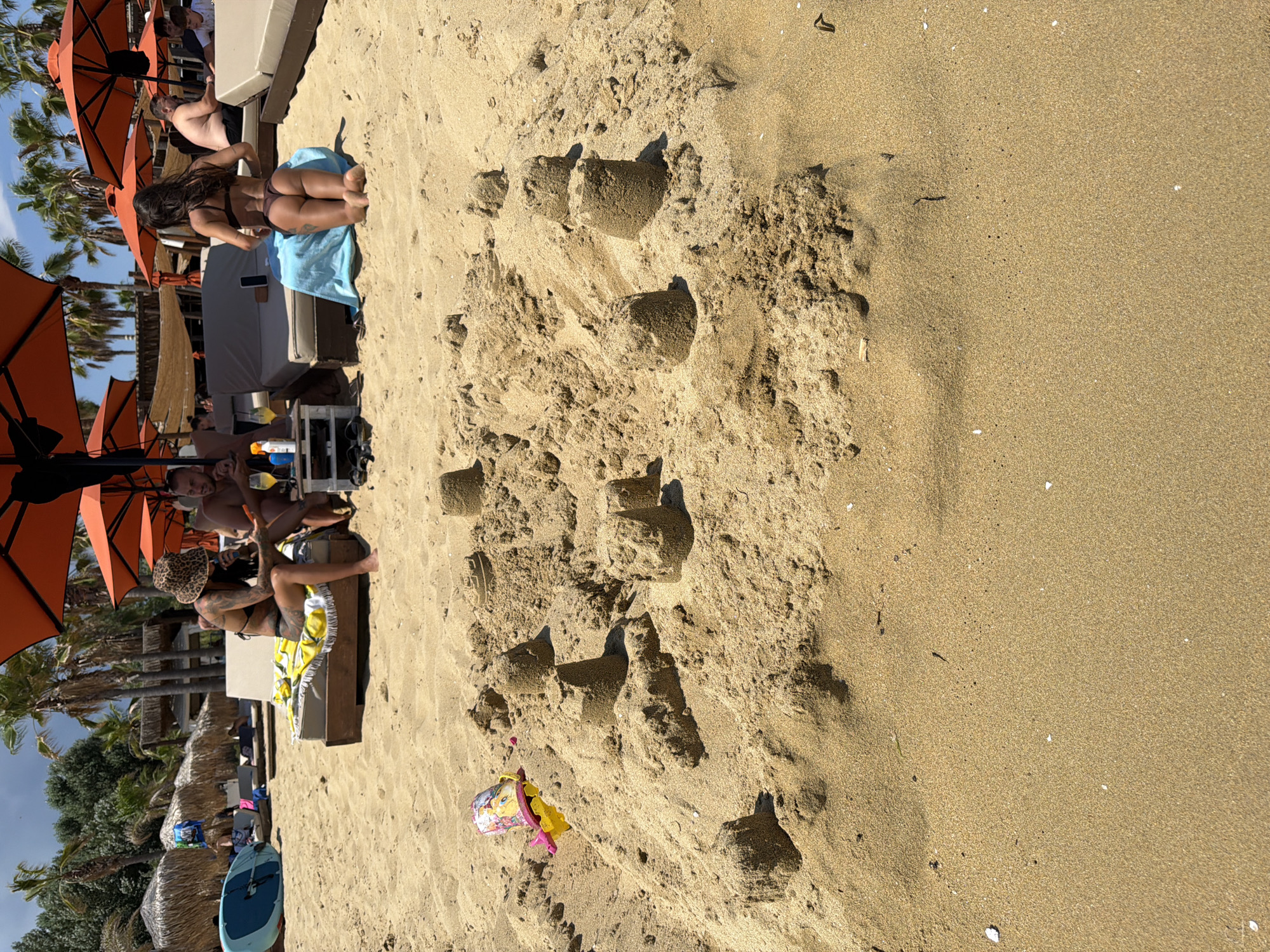 In Nea Peramos, Greece, a sun-drenched beach scene unfolds. In the foreground, an array of sandcastles, some meticulously crafted and others partially collapsed, occupy the sandy expanse. A child's yellow and pink toy bucket, adorned with cartoon characters, lies on its side amidst the sandy structures.
Further back, three individuals recline on lounge chairs positioned under large orange umbrellas. One woman, wearing a leopard-print bucket hat and a black bikini, is seated on a lounge chair, holding a phone and a drink. Beside her, a man, shirtless and with tattoos, also sits on a lounge chair, holding a beverage. Behind them, another woman in a dark bikini kneels on a blue towel on a lounge chair, her back to the camera. Other beachgoers and lounge chairs are visible in the background, along with palm trees and thatched-roof structures, suggesting a resort or beach club environment. The sea and sky are not prominently featured.