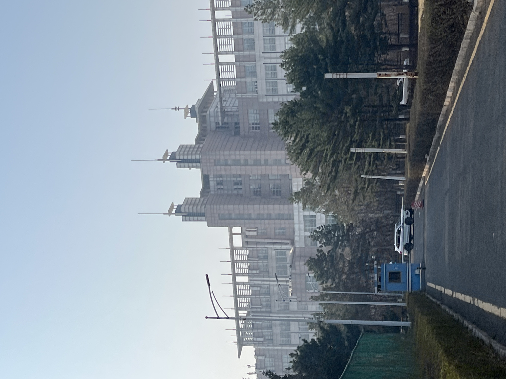This daytime image captures an urban or suburban street scene in Changchun City, China, viewed from a slightly elevated or angled perspective. On the right, a paved road or pathway, marked with a distinct yellow line, runs alongside a curb and a low hedge. Several tall, light-colored posts, likely lampposts, are evenly spaced along the road.
In the mid-ground, a white car is parked next to a small, blue, rectangular booth-like structure with a window, possibly a guard post or utility box. Behind the road and structures, a line of dense green trees and bushes forms a natural divider.
The background is dominated by several multi-story buildings with modern architectural designs, featuring numerous windows and complex structures on their upper levels, including decorative spires or antennas. Some parts of these buildings appear to be either under construction or have exposed frameworks, suggesting ongoing development. The buildings exhibit a slightly hazy appearance, possibly due to atmospheric conditions or distance.
The sky is a clear, light blue, occupying the upper left quadrant of the image. A distinctive streetlamp with a dark, curved arm is visible in the mid-left. The scene is quiet and still, with no discernible people or active events taking place. The fair weather and green foliage suggest a season other than winter. No visible text is present in the image.