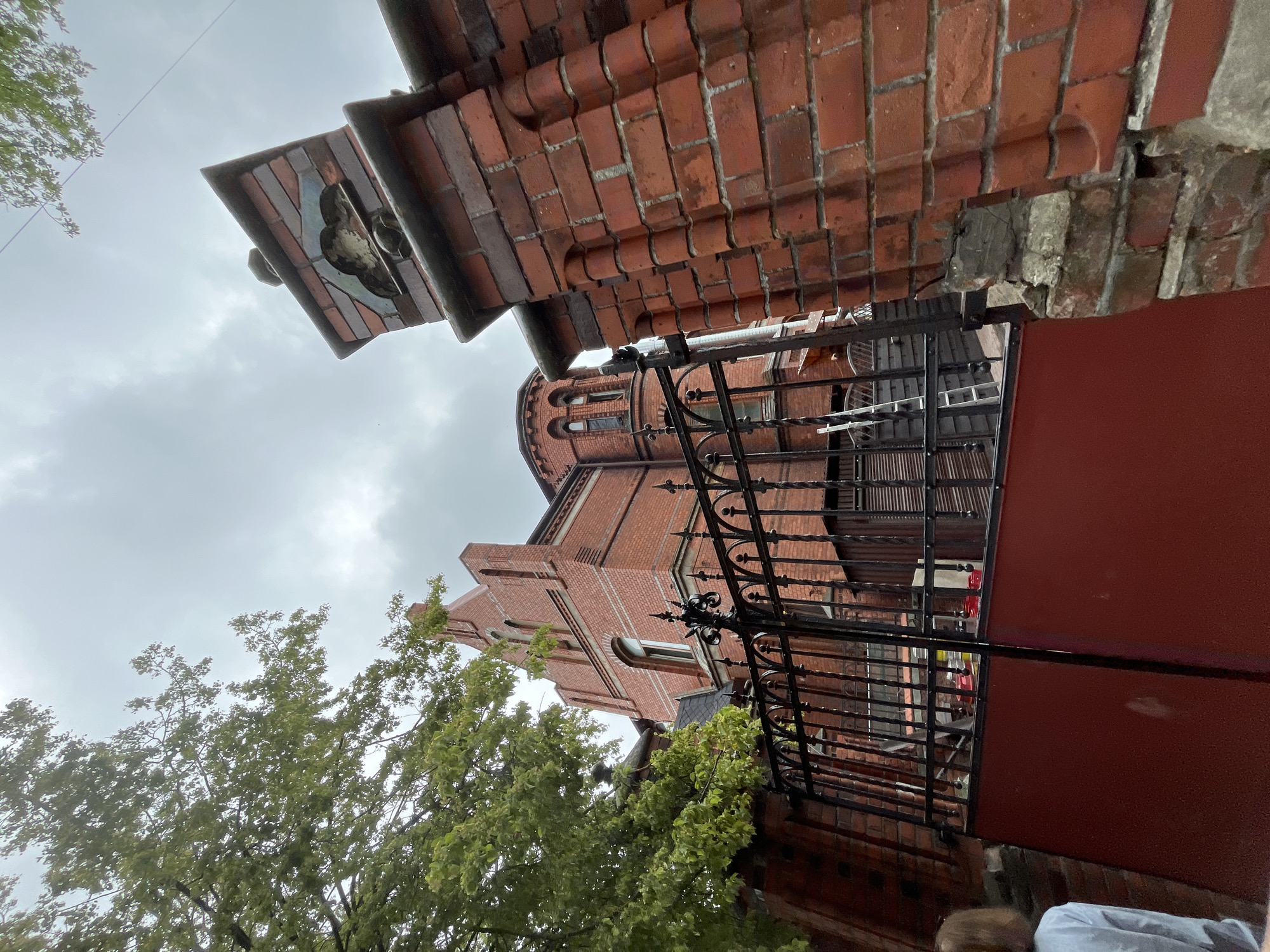 A brick fence with a decorative element on top and a wrought iron gate partially open in the foreground. Behind the fence, a large brick building with arched windows and a pointed roof is visible. A ladder leans against the building. A tree with green leaves is on the left. The sky is overcast. This is the building at 11 Pushkin Street in Zelenogradsk, Russia.
