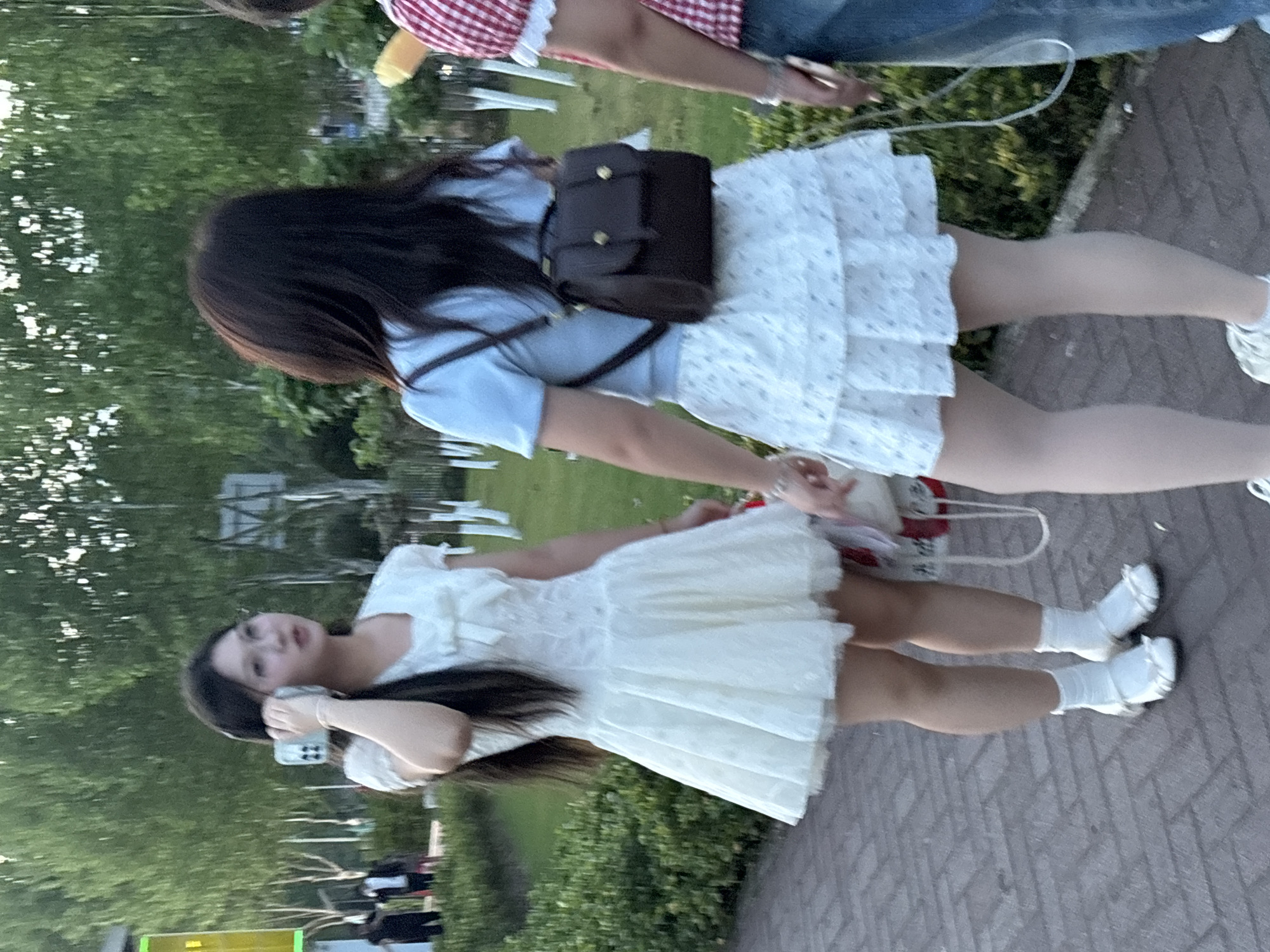 The image shows three young women walking on a paved path in a park-like setting. One woman wears a white dress and white socks with white shoes, and is holding a phone. Another wears a light blue top and a white skirt with small floral patterns, and a brown backpack. The third wears a red and white plaid top and jeans. The location is likely Jiangjin, China, though without additional information, confirmation is impossible. The background shows green trees and vegetation. There are white fence posts along the path.