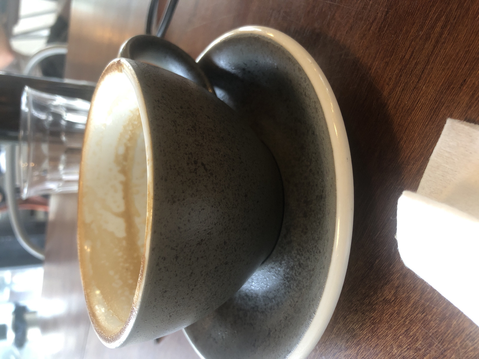 A close-up shot focuses on a gray ceramic coffee cup and saucer on a wooden table. The cup, which appears to be empty or mostly empty, has a light brown residue clinging to the inside walls, suggesting it recently contained coffee. A white napkin is partially visible in the foreground. In the background, a clear glass with water and another blurry glass are present, along with indistinct figures and furniture, indicating the setting is likely a café or restaurant. The lighting is soft, with natural light filtering in from the left, creating shadows and highlights. The image does not provide any specific clues to locate it in Đà Nẵng, Vietnam.
