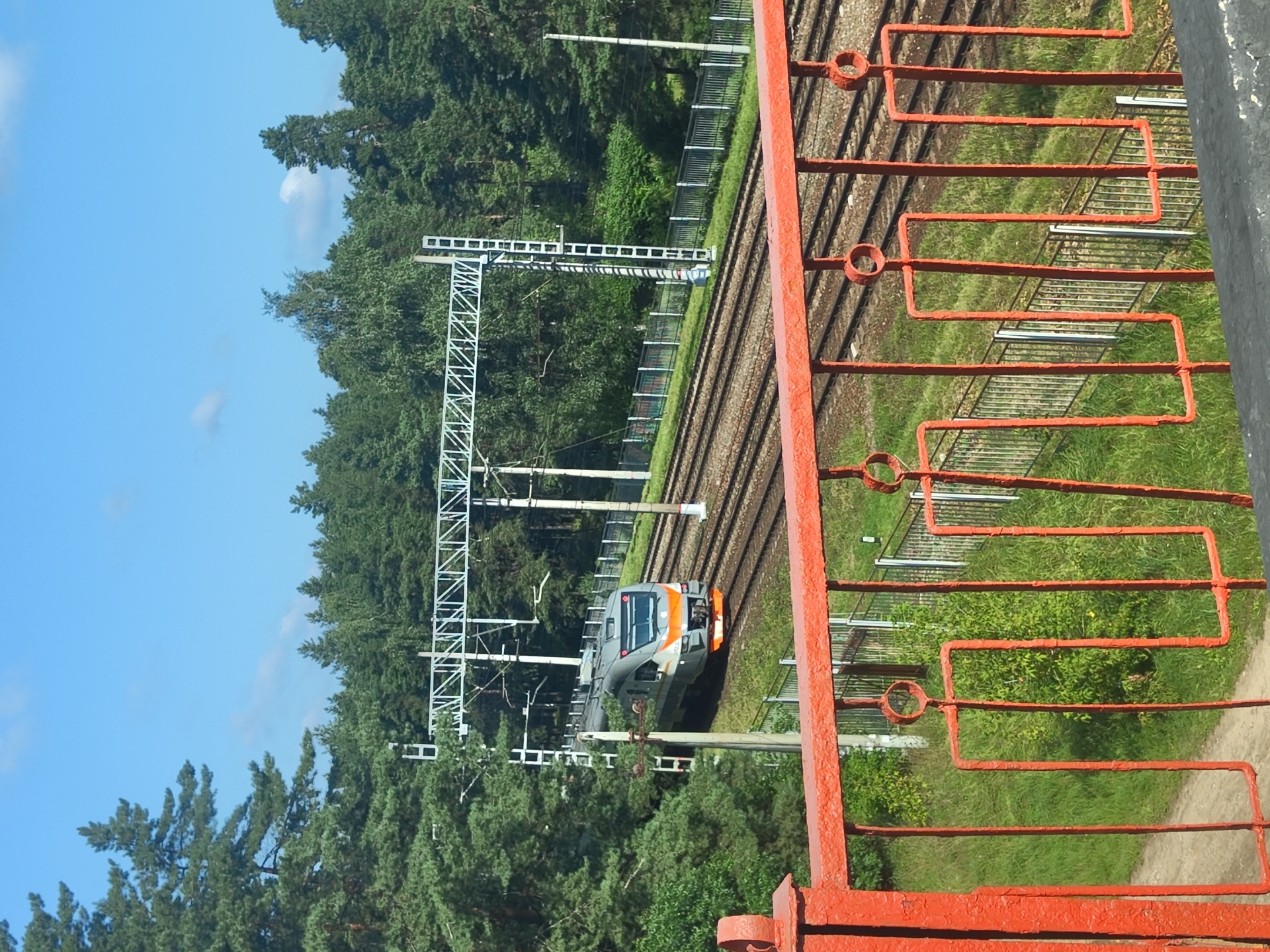 An outdoor, eye-level, slightly angled medium shot captures a modern, gray and orange train moving on a railroad track. The train is on the right side of the frame, heading towards the viewer. The railroad track runs diagonally from the bottom right to the top left. The scene is framed by an orange metal railing in the foreground. Lush green trees and dense forest form the backdrop, with a bright blue sky with a few scattered white clouds above. Overhead, a white metal structure with electrical wires is visible above the train, indicating an electrified railway line. A white metal fence runs along the right side of the tracks. The image appears to be taken on a sunny day. While the prompt mentions Ramenskoye, Russia, there are no specific landmarks or identifying features within the image to confirm this exact location.