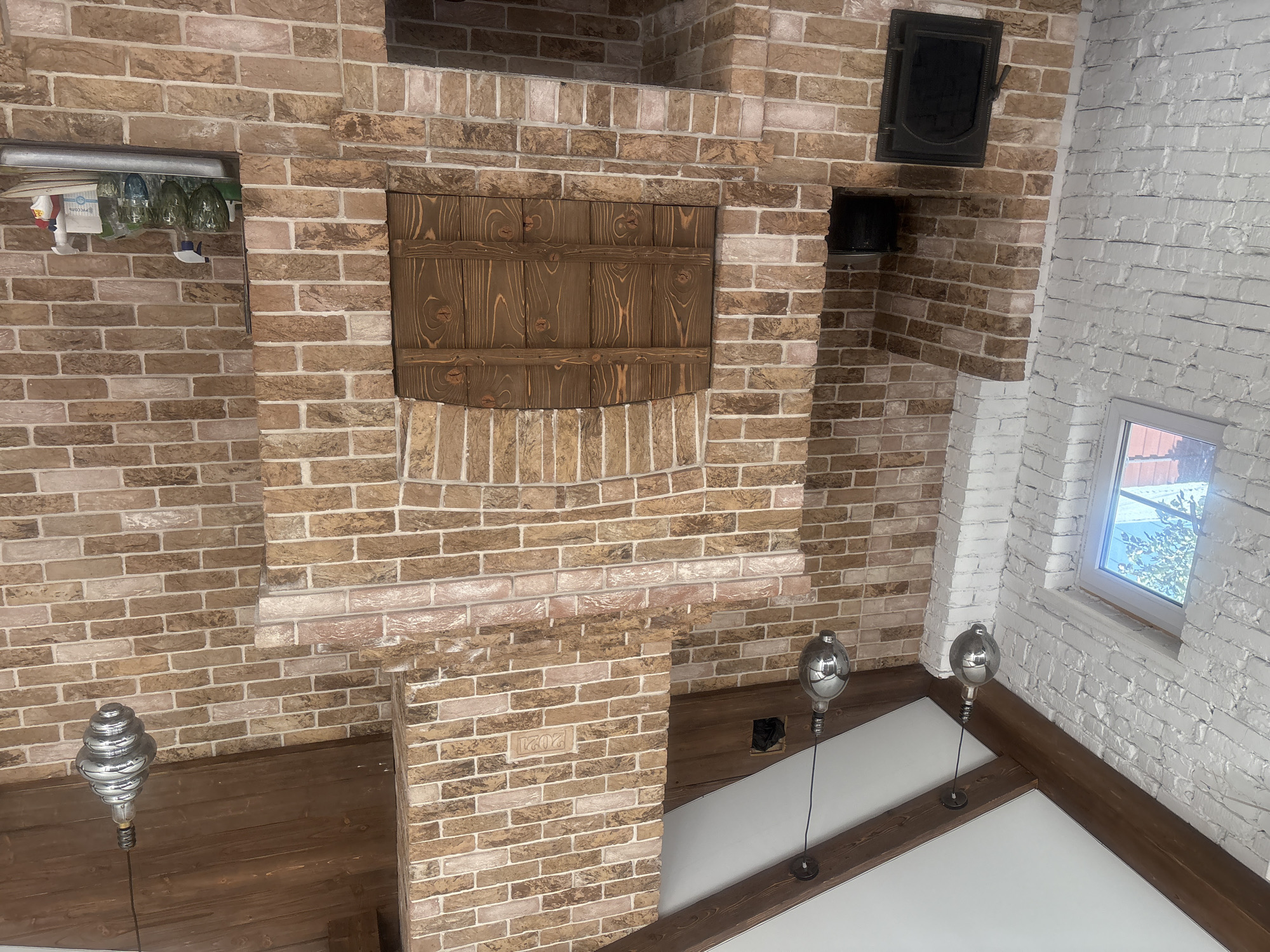 The image shows an interior view of a brick structure, possibly a covered patio or outdoor kitchen, with a dominant brick fireplace and oven. The fireplace is constructed with a mix of light and dark brown bricks, featuring an arched opening with wooden doors. To the left, a wall is partially visible, constructed with white-painted bricks, and includes a window with a view of trees and a red-tiled roof. The ceiling has exposed wooden beams and is white. Several industrial-style pendant light fixtures hang from the ceiling. On a shelf attached to the brick structure, there are several glasses, spray bottles, and cleaning supplies. A black metal door is set into the lower part of the brick structure. A black pot is visible inside an alcove above the metal door. The year "2021" is inscribed into one of the bricks on the fireplace. The scene is located in Georgiyevsk, Russia.
