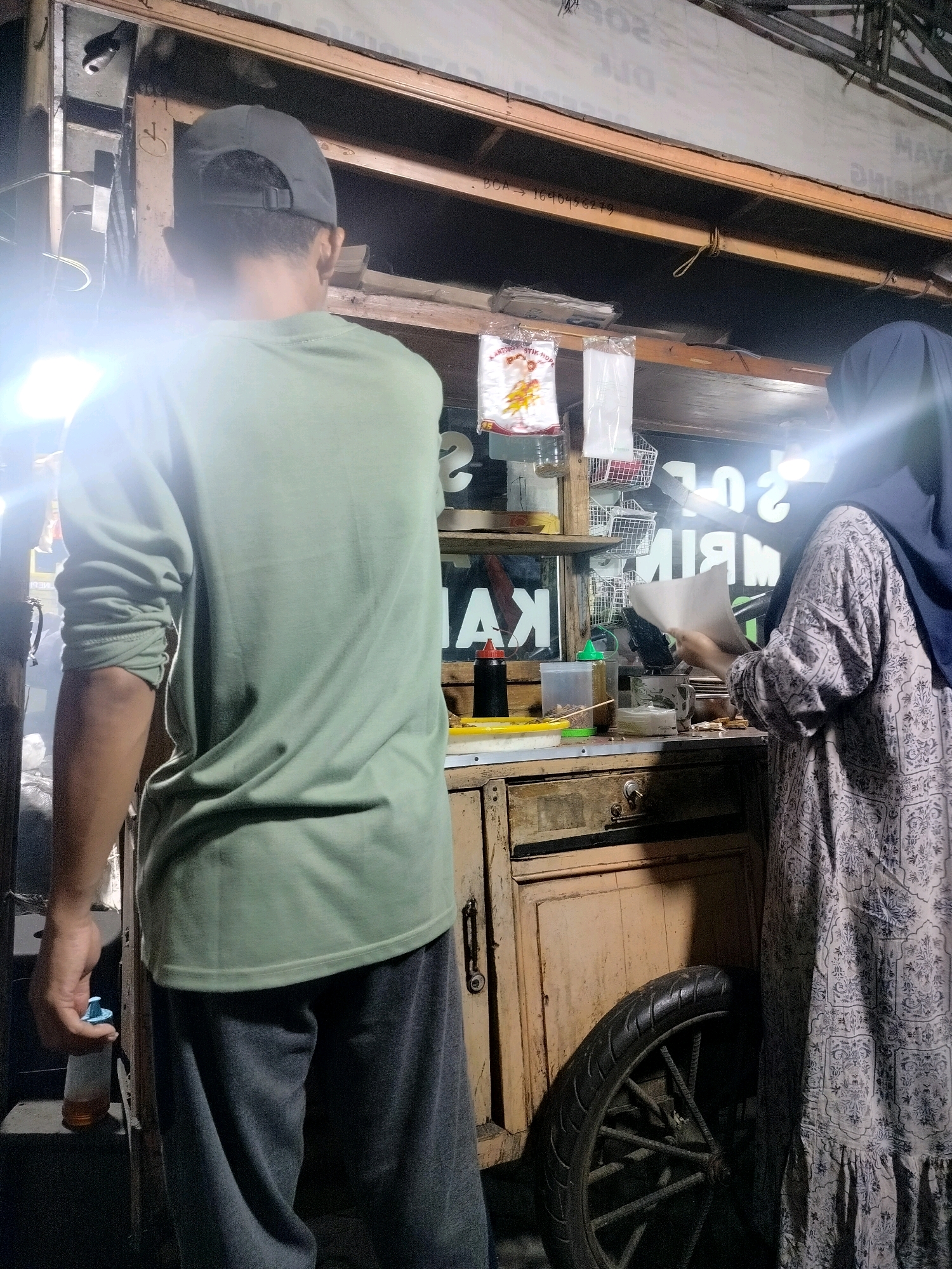 A man wearing a green long-sleeved shirt and a dark grey baseball cap stands with his back to the camera, facing a food cart. To his right, a woman wearing a dark blue hijab and a patterned grey dress is preparing food on the cart. The cart appears to be made of wood with some metal components. There are various items on the cart, including bottles of condiments, containers of food, and bags of what appear to be packaging materials. A wheel is visible on the lower right side of the cart. The scene is dimly lit, with artificial light sources contributing to the overall illumination. The image appears to be taken in an outdoor urban setting, possibly in Bekasi, Indonesia, given the context.
