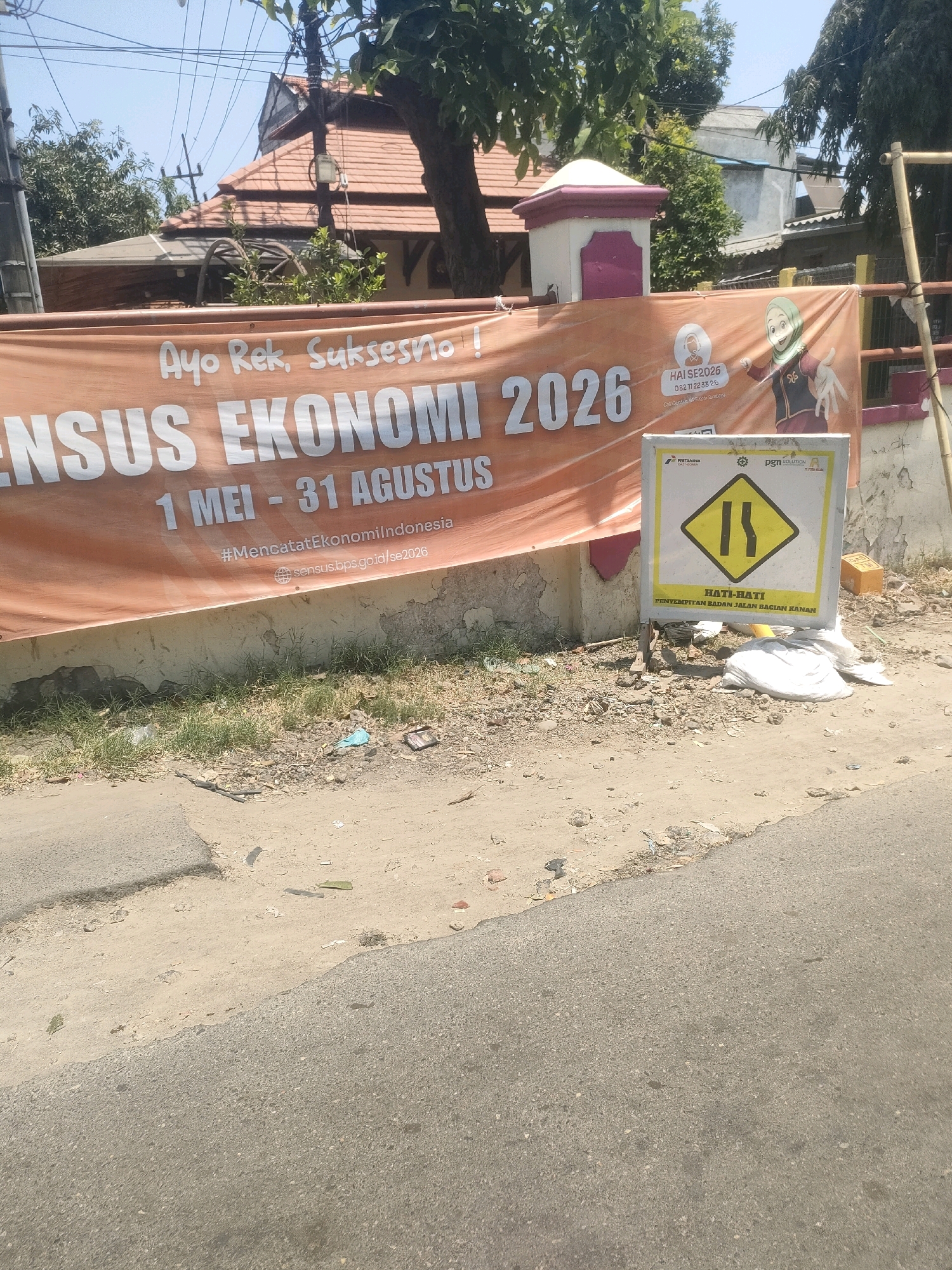 An outdoor, eye-level, medium shot captures a street scene in Surabaya, Indonesia, on a bright, sunny day. In the foreground, a dusty, uneven road surface transitions to a sidewalk area with scattered debris. Dominating the mid-ground is a large, orange banner advertising the "SENSUS EKONOMI 2026" (Economic Census 2026), with the dates "1 MEI - 31 AGUSTUS" (May 1 - August 31) also displayed. The banner features text in Indonesian, including "#MencatatEkonomiIndonesia" and a website address. To the right of the banner, a yellow diamond-shaped traffic sign with black markings warns of a narrowing road, indicating a lane reduction on the right. The sign also displays text in Indonesian: "HATI-HATI PENYEMPITAN JALAN BAGIAN KANAN" (Caution Narrowing Road Right Side). Adjacent to the sign are various items including white plastic bags, a yellow cardboard box, and debris. In the background, a building with a red-tiled roof and a white pillar with purple accents is visible, along with trees and overhead electrical wires. The scene appears to be a public street.
