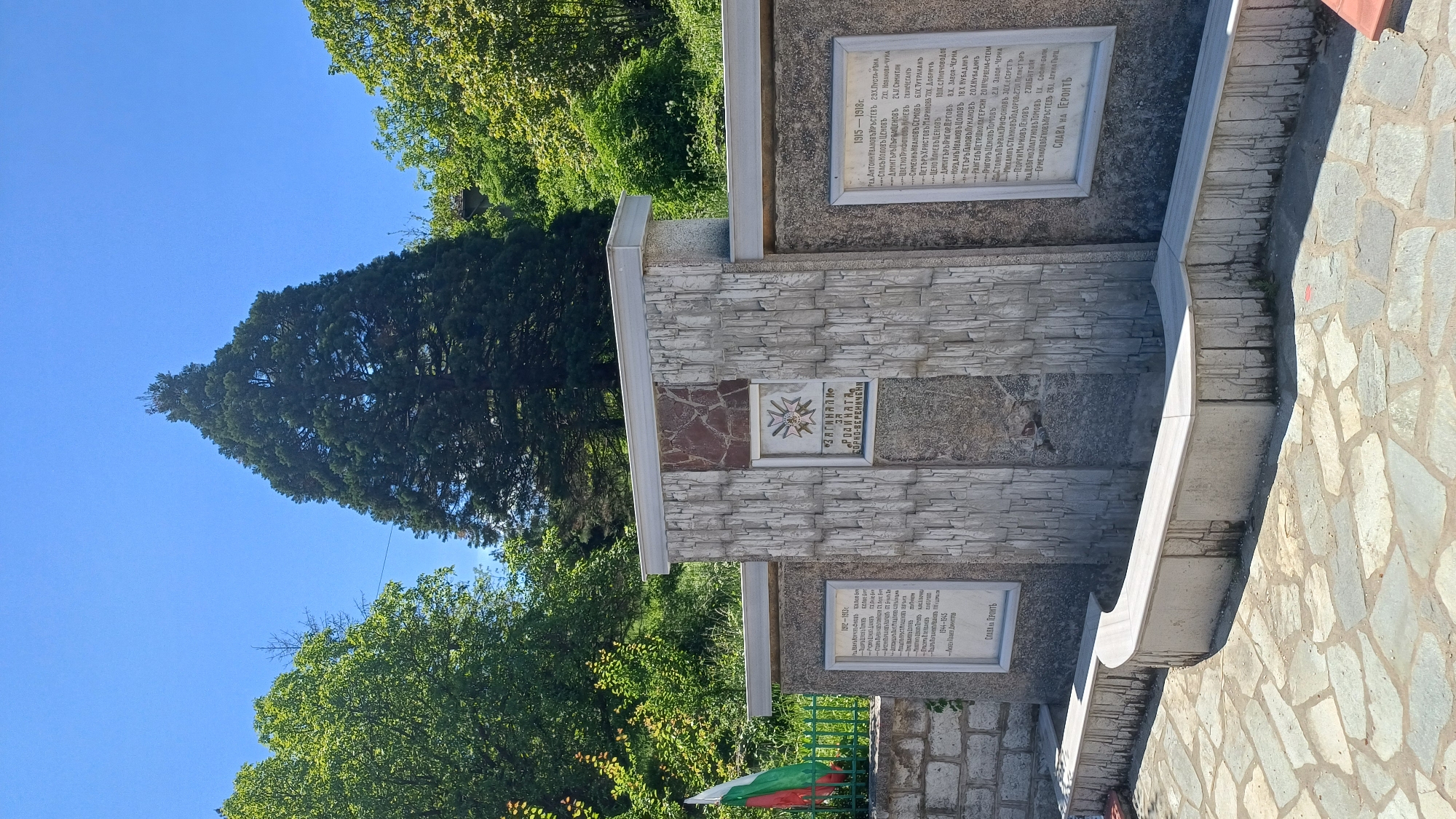 The image shows a stone monument in Gorna Verenitsa, Bulgaria. The monument is made of light gray stone blocks and contains three plaques with text. The top plaque lists names from 1915-1918, and the bottom plaque lists names from 1941-1945. The central plaque features an emblem and the words "Загинали за родината Горна Вереница" (Fallen for the homeland Gorna Verenitsa). The Bulgarian flag is visible next to the monument. Behind the monument are trees and a blue sky. The monument is located next to a stone-paved area.
