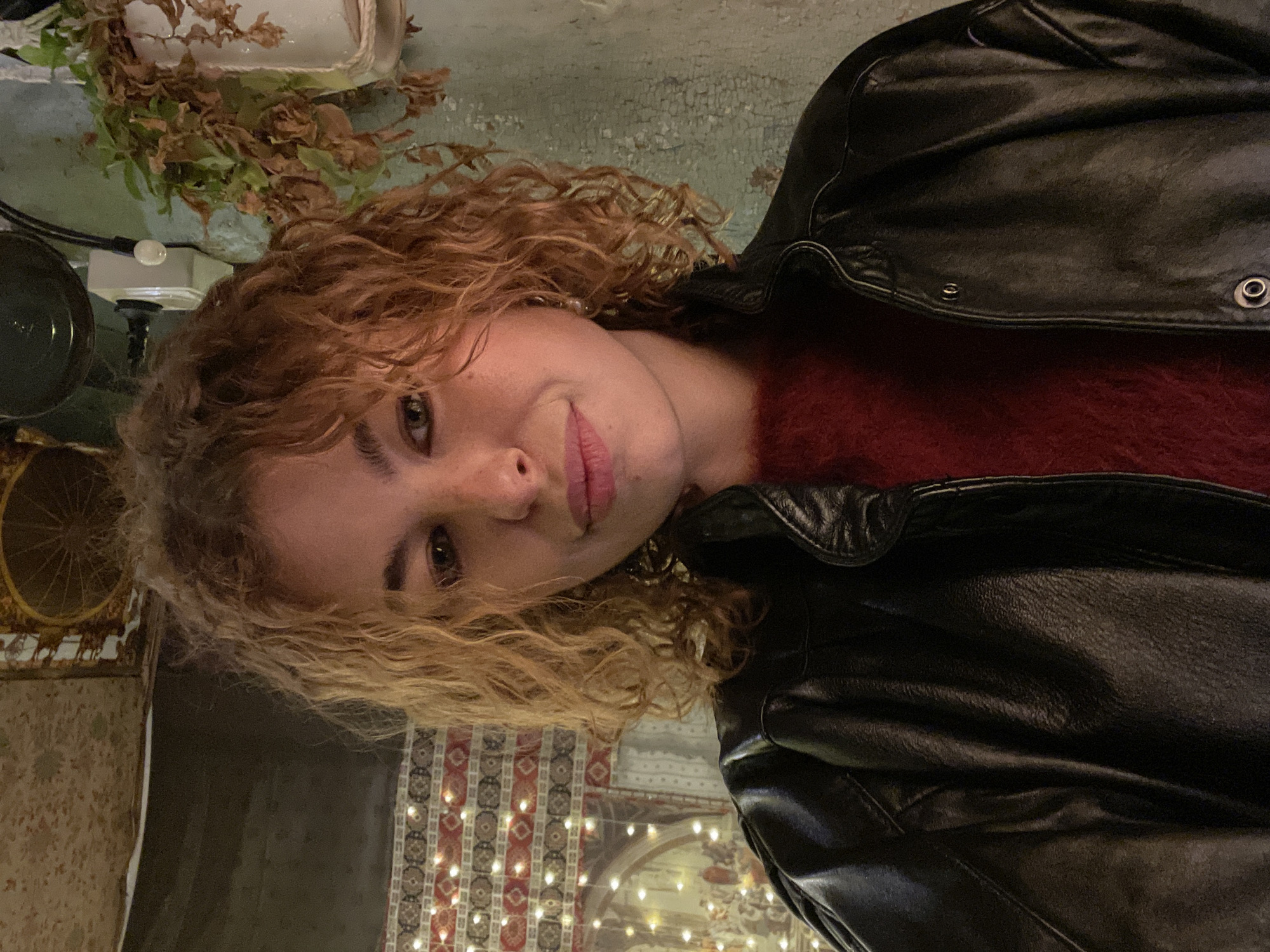 The image depicts a fair-skinned individual with curly red-blonde hair, wearing a red fuzzy sweater and a black leather jacket. The location appears to be a restaurant interior, as indicated by the string lights, patterned wallpaper, and decorative elements. Based on the image detail, the setting is likely a restaurant or establishment in Tbilisi, Georgia, as the wallpaper design appears to reflect the city’s distinctive aesthetic. The individual is positioned at an angle, facing the camera with a slight smile.