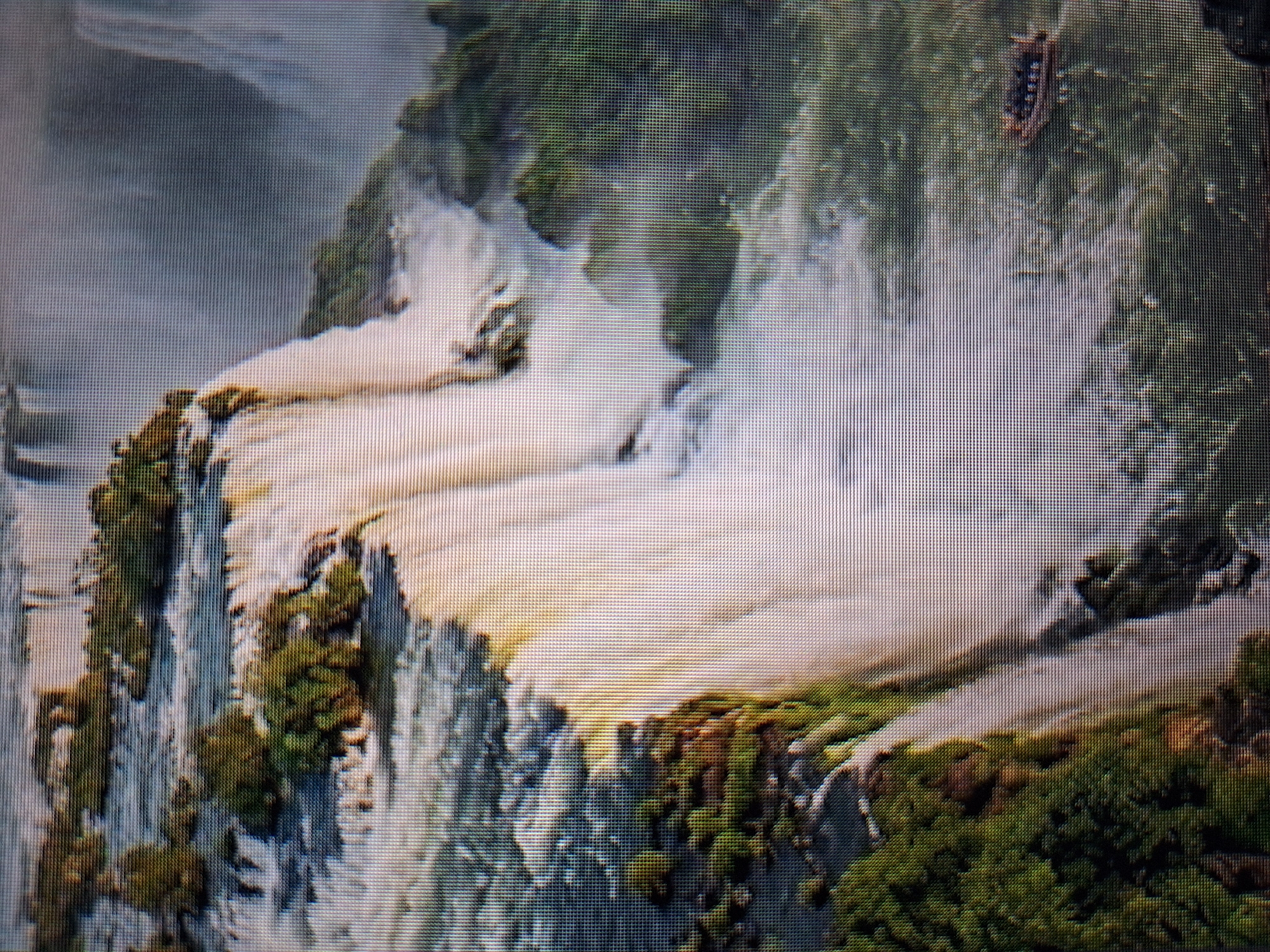The image shows a wide shot of a massive waterfall system with lush green vegetation on the surrounding cliffs. The water cascades down in multiple tiers, creating a powerful and awe-inspiring spectacle. In the lower right portion of the image, a small boat carrying several passengers is visible navigating the water near the base of the falls. The overall scene depicts a natural wonder, likely a tourist attraction, characterized by its immense scale and the raw power of nature. The image is a photograph taken from an elevated perspective, offering a comprehensive view of the waterfall and its surroundings. The context of Varna, Bulgaria is not apparent in the image itself.
