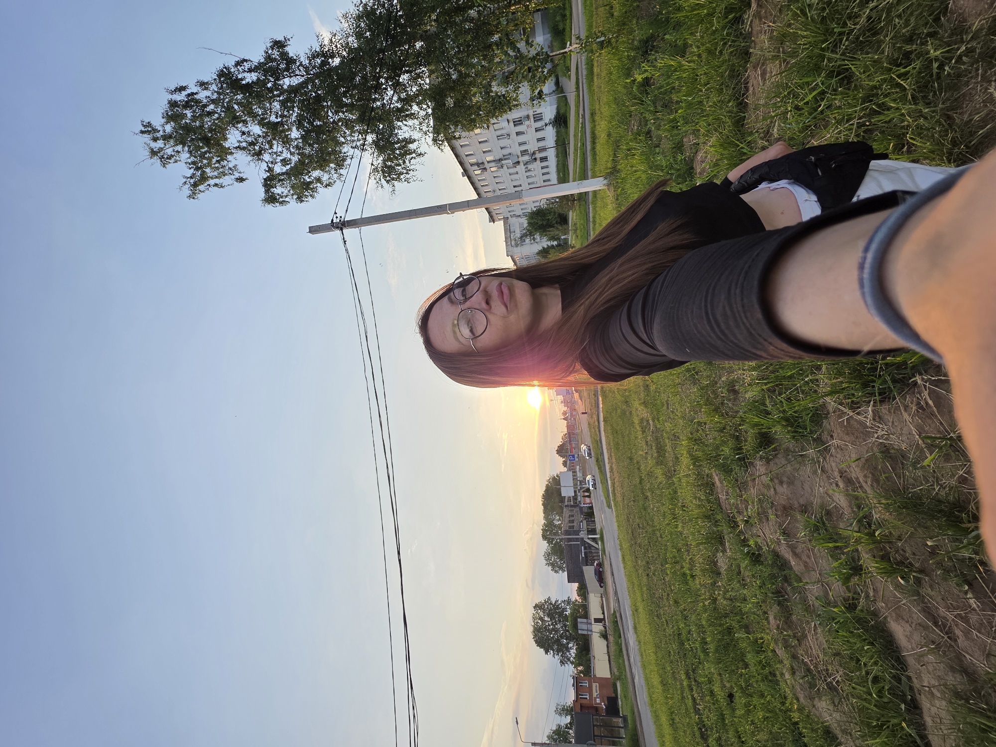 A woman is taking a selfie outdoors in Chaikovsky, Russia. She is wearing a black long-sleeved top, white pants, and glasses. She has long brown hair and is looking at the camera with her lips pursed. The background shows a street with cars, buildings, trees, and a utility pole. The sun is setting, casting a warm glow on the scene. The overall environment appears to be a residential or commercial area on the outskirts of the city, with a grassy field in the foreground.