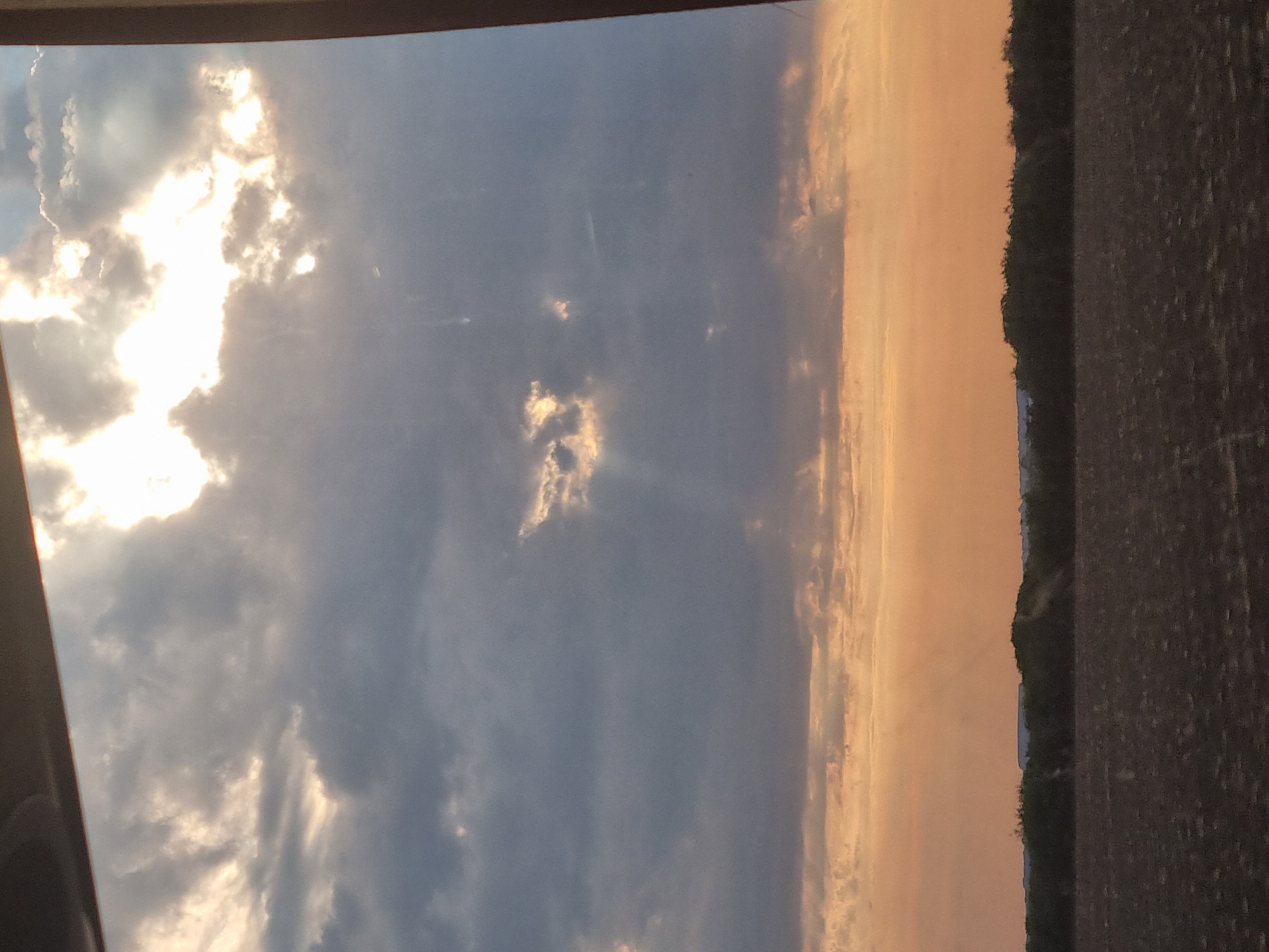The image depicts a sunset viewed from inside a vehicle, likely a car or bus, as indicated by the dark frame at the top and the right edge of the image suggesting a window or windshield. The sky is dominated by dramatic clouds, with the sun breaking through in the upper right quadrant, casting bright rays. The lower part of the sky transitions into a warm, orange glow. Below the sky, a dark line of trees forms a silhouette against the horizon. The foreground consists of a dark, textured field, possibly farmland. The scene is a natural landscape at dusk, with no visible people or specific urban features that would pinpoint a location within Cefa, Romania.