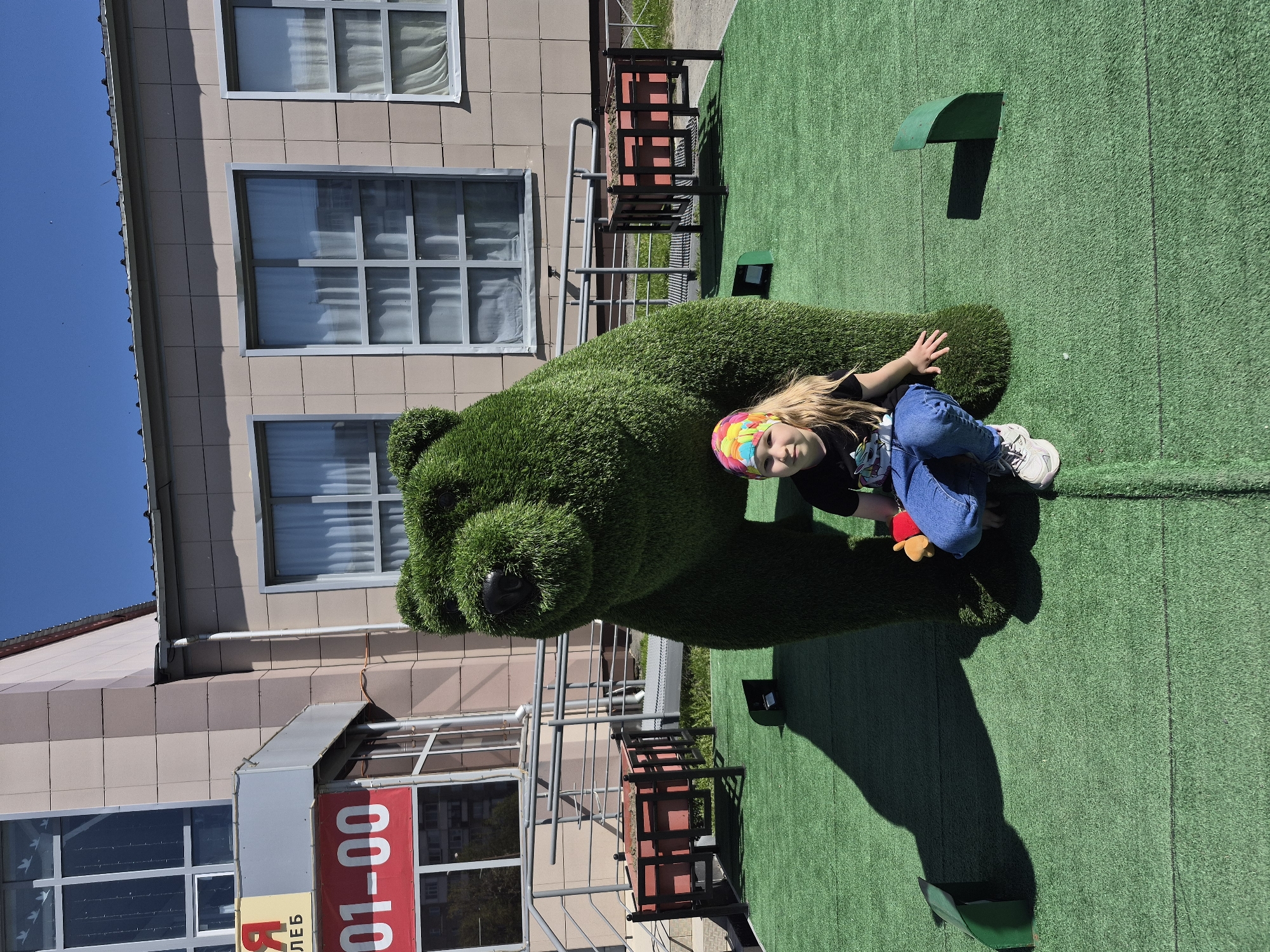 A young girl is sitting on an artificial grass bear figure, posing for a photograph on a bright day. She wears a multicolored headband, a dark t-shirt, blue jeans, and white sneakers. The bear is sculpted from green artificial grass.
The background includes a multi-story building with beige-colored tiled exterior walls and rectangular windows. A sign with the numbers "01-00" is visible at the bottom left of the frame. The surrounding surface is covered in artificial green grass.
Based on the available information, this photograph was taken in the city of Tobolsk, Russia.