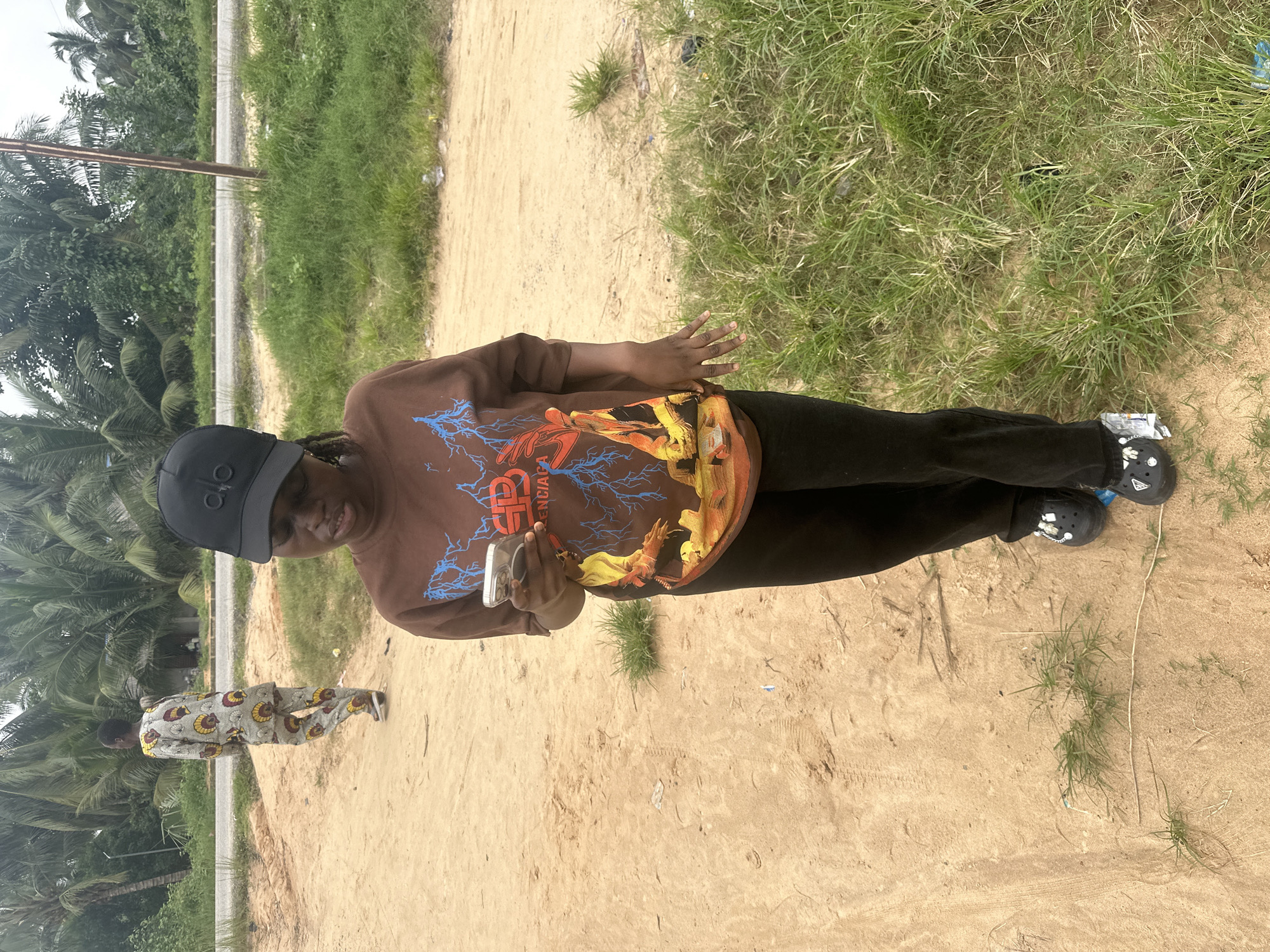 The image shows a young woman standing on a dirt path, looking at her cell phone. She is wearing a brown sweatshirt with a graphic design, black pants, and black Crocs. She has on a black baseball cap. In the background, another person in a patterned outfit walks away from the camera. The setting appears to be rural, with palm trees and other vegetation visible. A paved road runs in the distance. The location is identified as Ganyingbo, Nigeria.