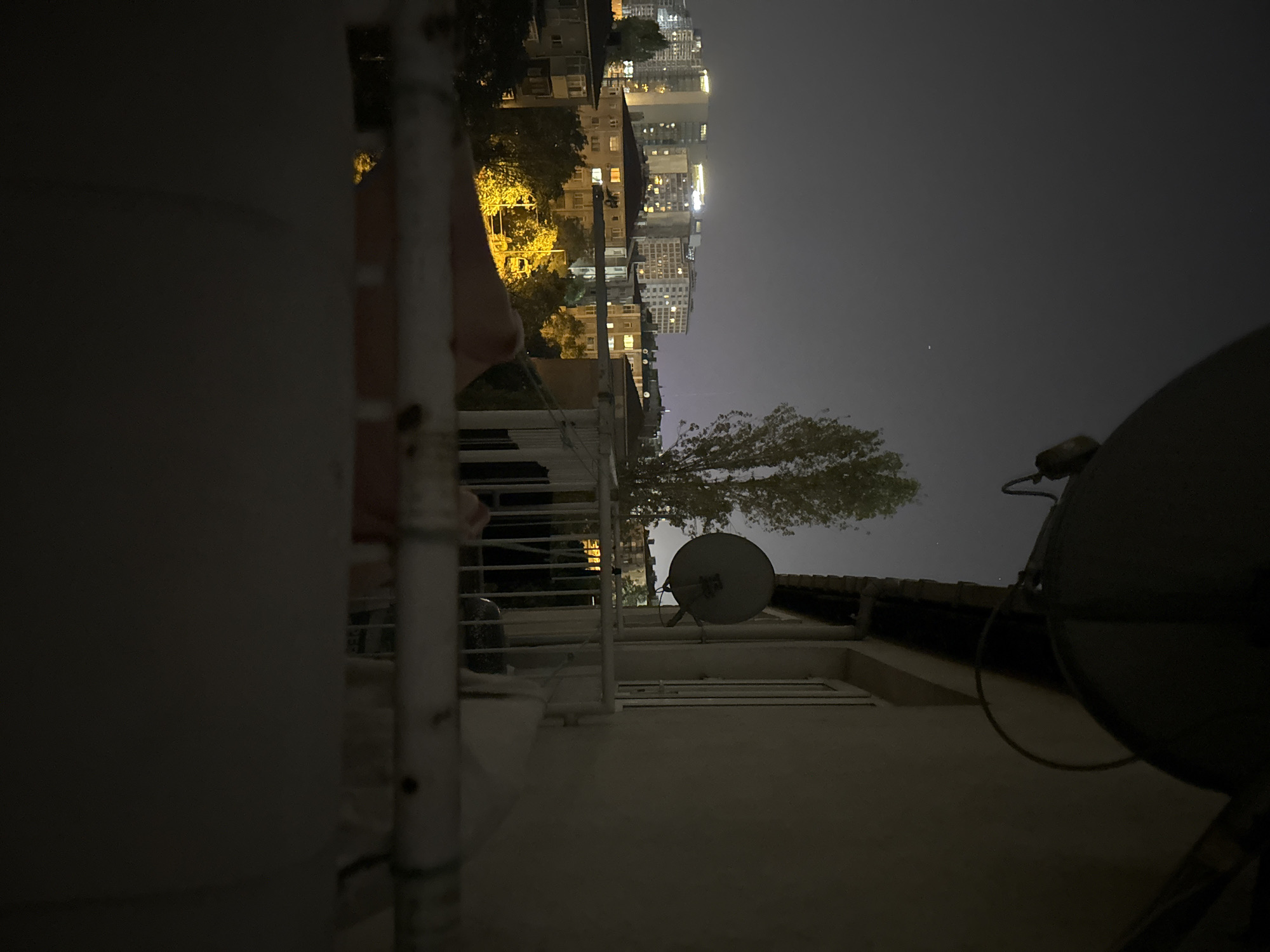 The image shows a night-time cityscape in Güngören, Turkey, viewed from a balcony. In the foreground, the edge of a balcony railing is visible, with a blurred white pipe running horizontally across the lower portion of the frame. Above the railing, a large satellite dish is mounted on the side of a building, with another smaller satellite dish on the balcony. Beyond the balcony, a dense urban area with numerous apartment buildings and trees is illuminated by artificial lights. The sky is dark and hazy, typical of a city at night. A single faint star is visible in the upper left quadrant of the sky. The overall impression is of a typical residential neighborhood in a Turkish city at night.