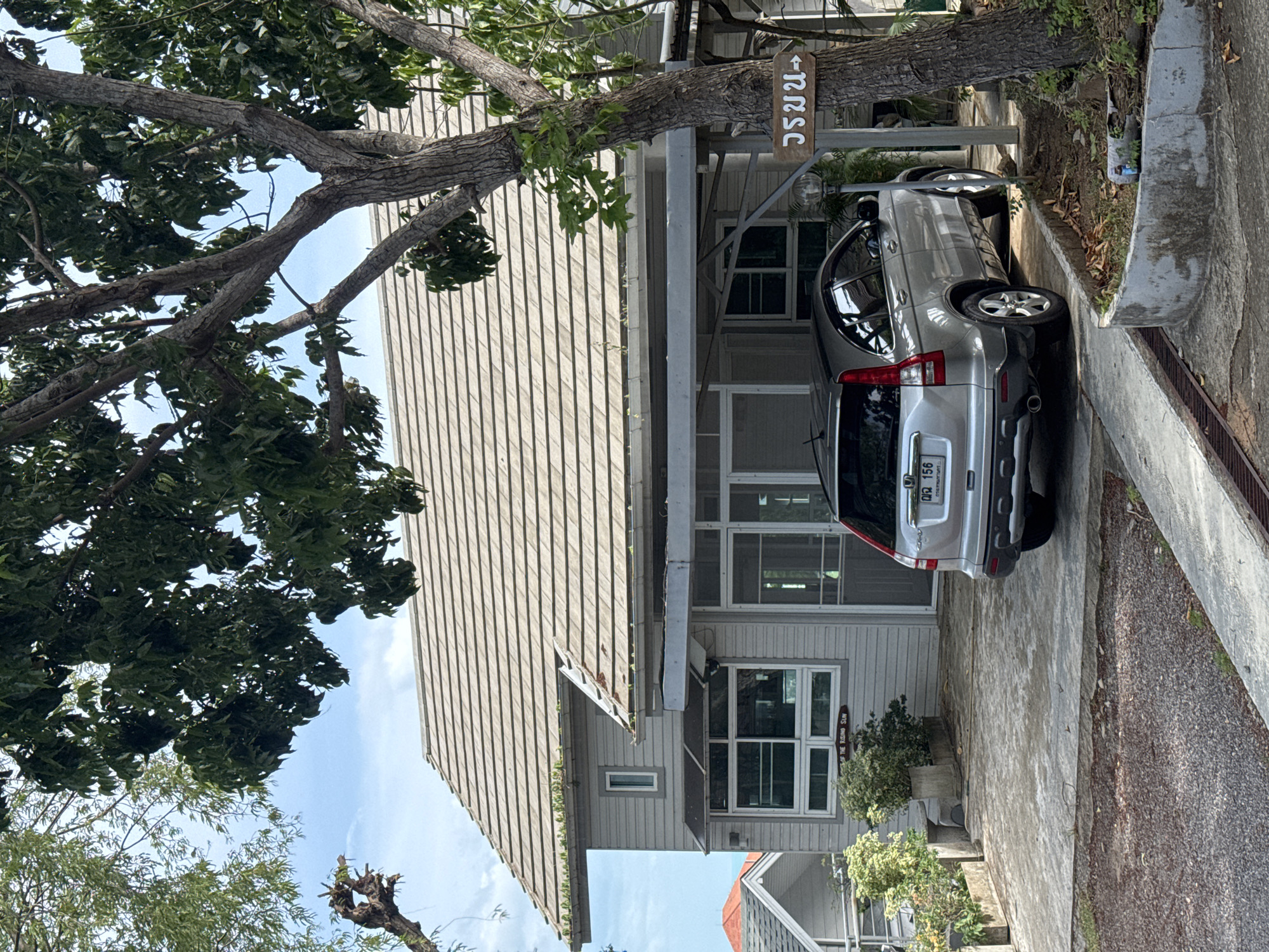 In the city of Ko Samui District, Thailand, a silver Honda CR-V is parked on a concrete driveway in front of a light gray house with a beige tiled roof. A wooden sign with Thai writing points to the right. Trees with green leaves surround the property. The sky is cloudy.