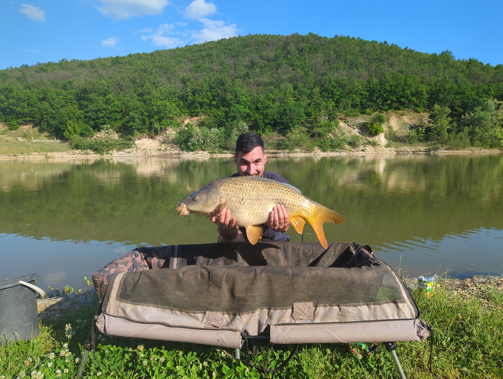 A man is holding a large carp in front of a lake. The man has short dark hair and is smiling. He is standing behind a fishing unhooking mat. The lake is surrounded by green trees and hills. The sky is blue with a few clouds. The location is Slavovitsa, Bulgaria.