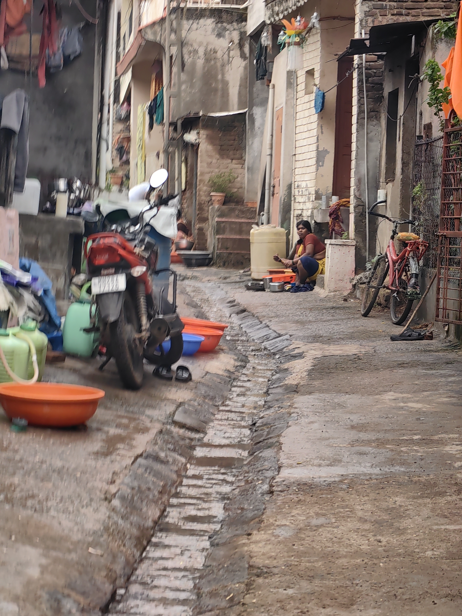In Mhaiswadi, India, a narrow, wet alleyway is depicted, suggesting recent rain. The ground is paved with stones and concrete, with a visible drainage channel running along the left side. On the left, a red motorcycle is parked, leaning against a wall covered in hanging clothes. Nearby, several plastic containers, one large and orange, are scattered. In the background, on the right side of the alley, a woman wearing a maroon shirt and blue shorts is sitting on the ground, interacting with cooking utensils and containers. Adjacent to her, a red bicycle is parked. The buildings lining the alley are multi-story structures with weathered facades and exposed brickwork. Laundry hangs from windows and balconies, indicating residential activity. The overall atmosphere is one of everyday life in a densely populated urban environment.