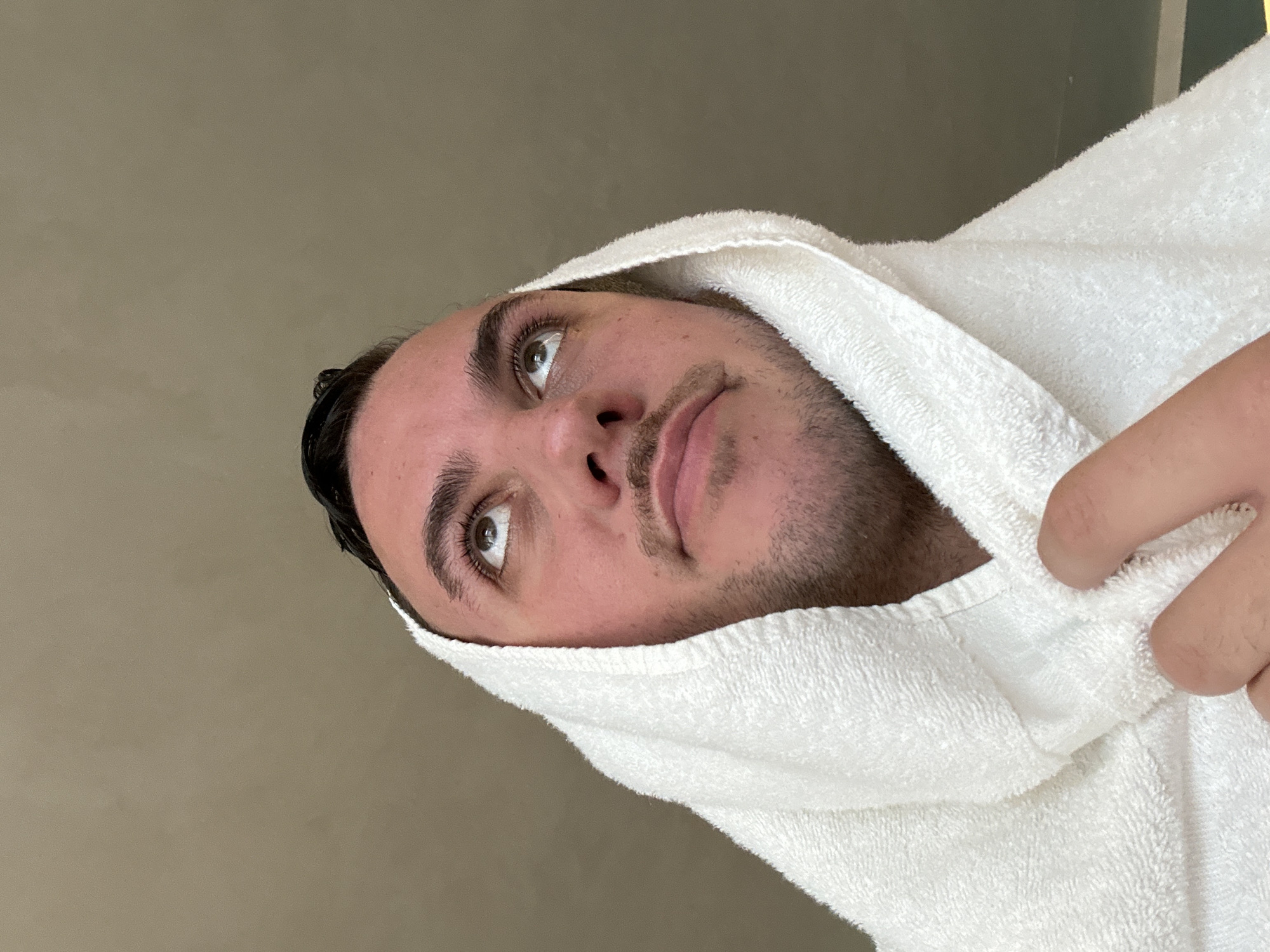 A close-up shot focuses on a person's face framed by a white towel draped over their head, possibly after a shower. The individual has dark hair, distinct eyebrows, brown eyes looking upward, a moustache and stubble. Their skin appears light in tone. A hand is also visible, holding the towel in place. The background is a plain, light-colored wall. The scene appears to be indoors and the provided image does not include any information to provide a location in the city of London, United Kingdom.