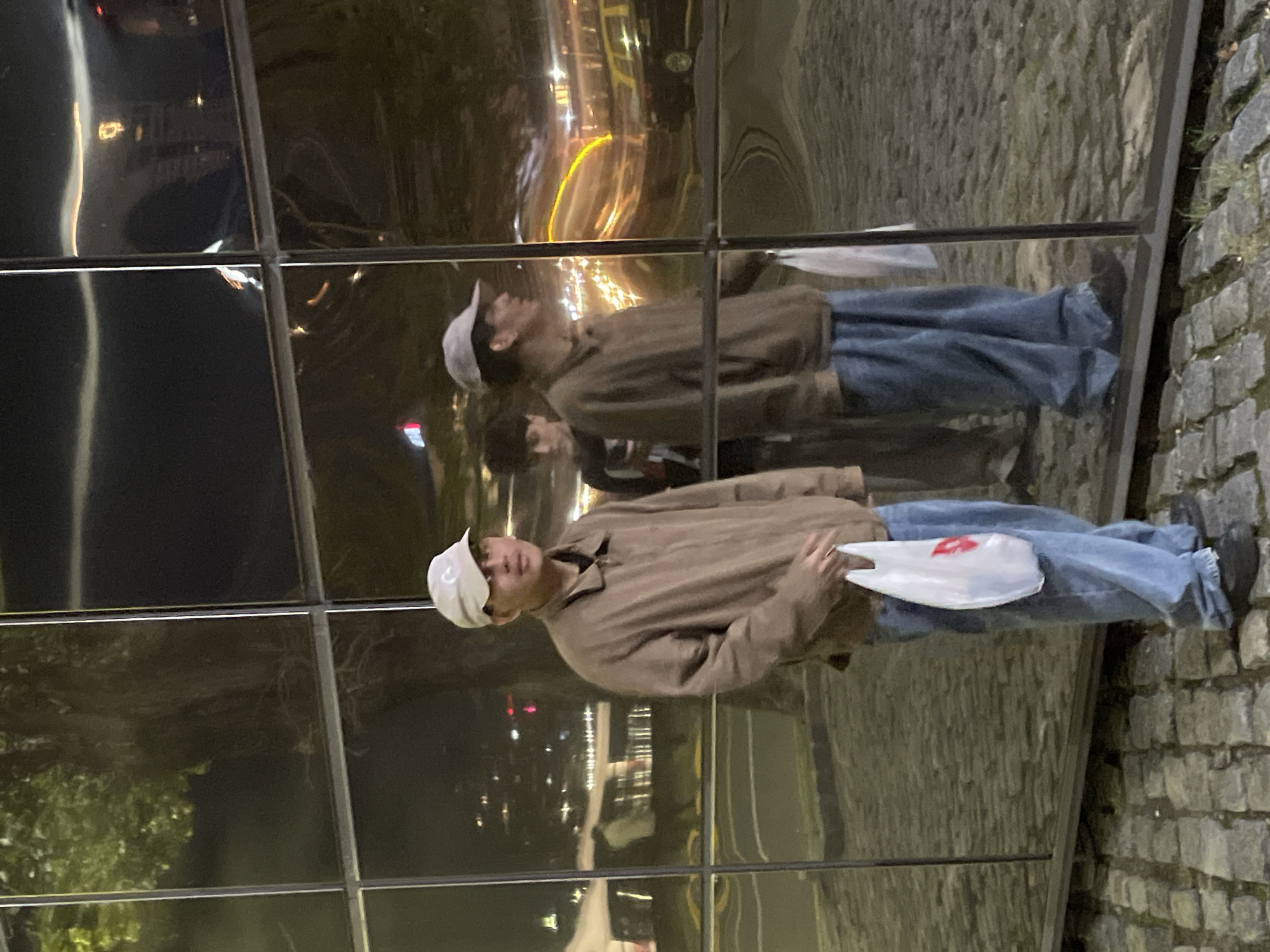 A man is reflected in a metallic wall. He is wearing a white cap, a tan jacket, and blue jeans. He is holding a white plastic bag. The reflection also includes street lights and trees. The wall is divided into square panels. The background appears to be a street. A stone wall is to the right. The location is likely in a city but the specific location of Buenos Aires, Argentina cannot be determined from the image.